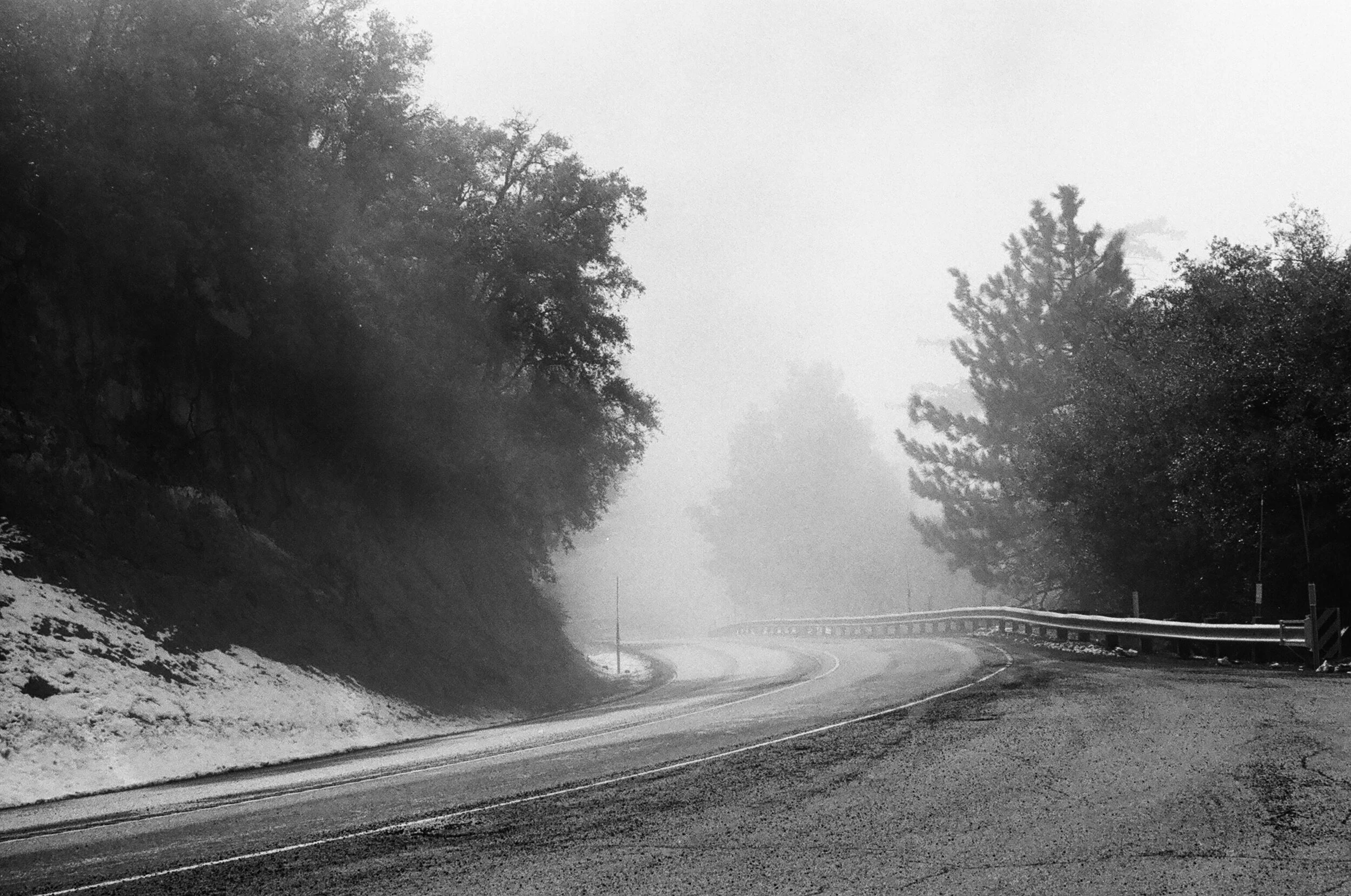 A foggy mountain road with trees on both sides and a guardrail on the right, with snow on the edges of the road.