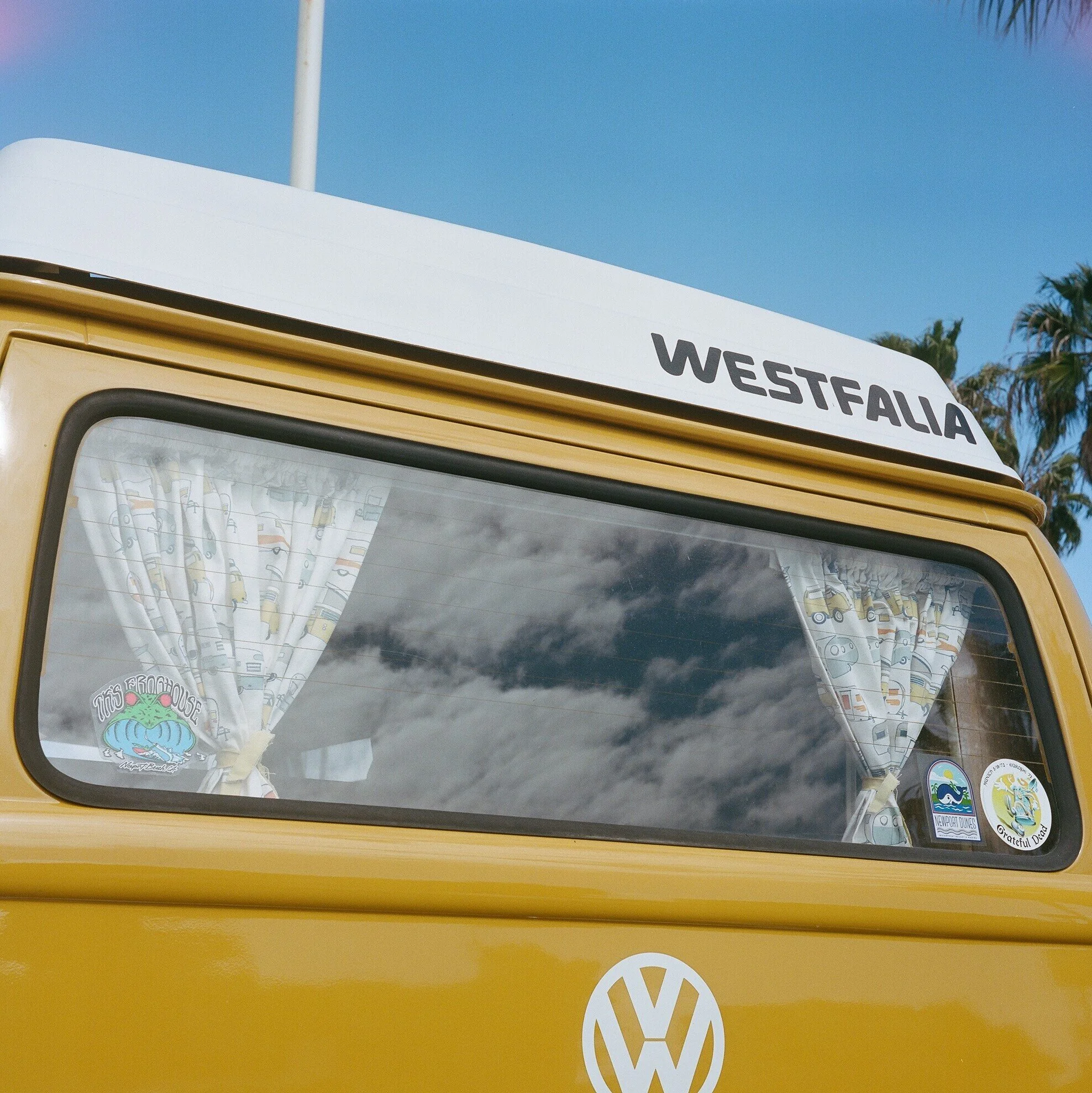 The front of a yellow Volkswagen van with a white roof and the word 'WESTFALIA' on a sign on top. The van has a large window with colorful curtains and several stickers, including one with the words 'The Argonauts' and a whale design. The background 