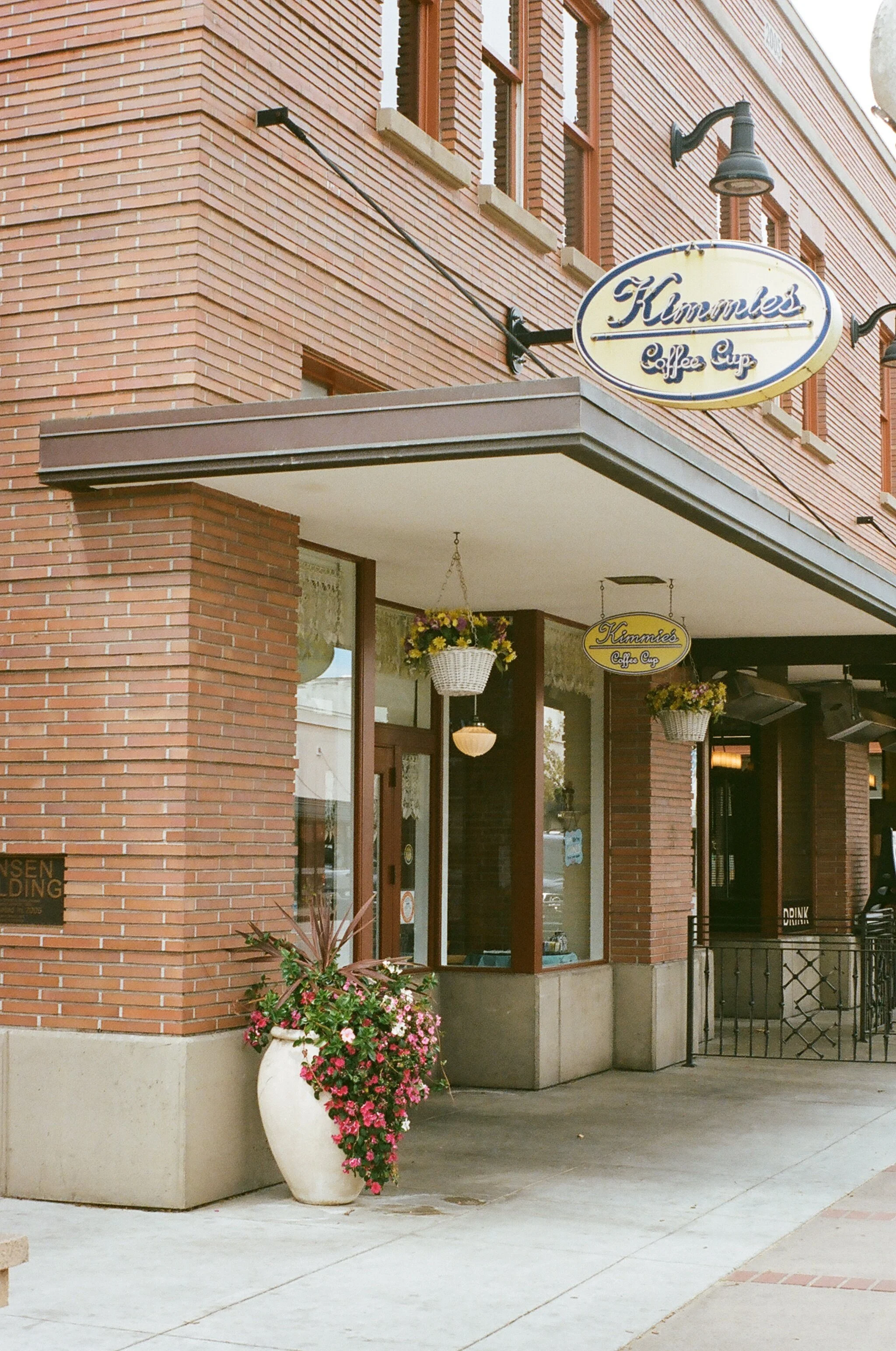 Exterior view of Kammie's Coffee Cup shop with a hanging sign, large flowerpot with pink and purple flowers, and hanging flower baskets.