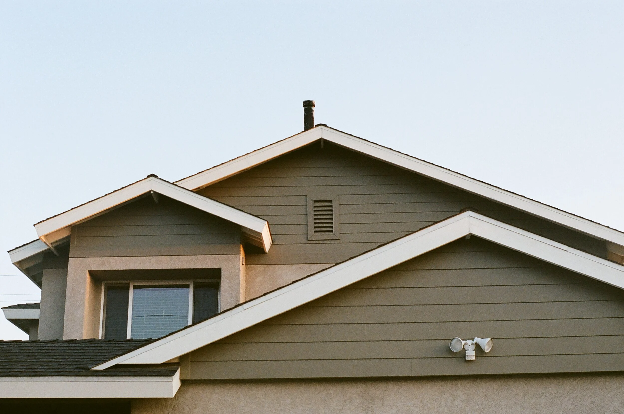 Close-up of the roof and exterior walls of a modern house with gray and beige siding, a small window, and a security camera.