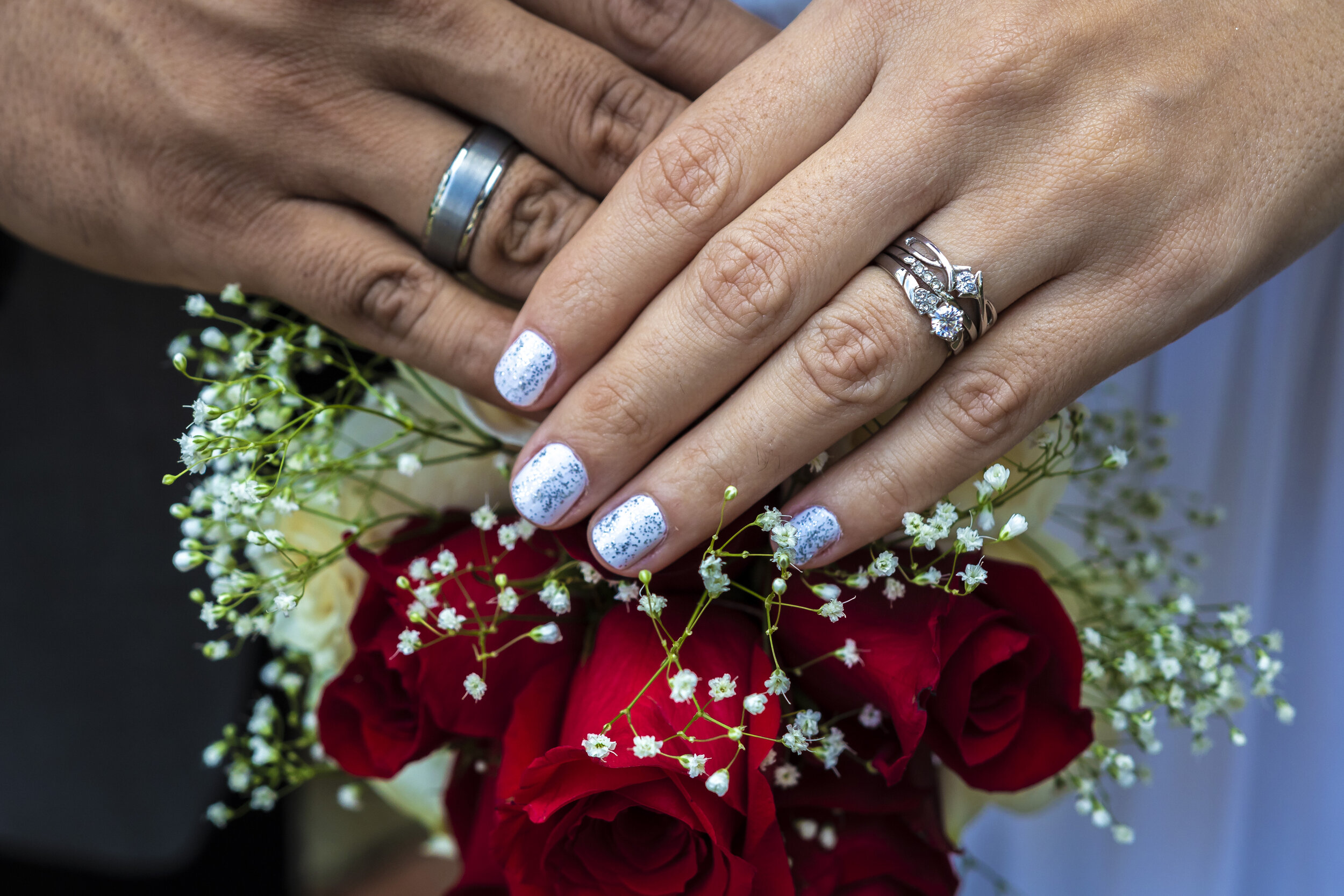 Close-up of two hands with wedding and engagement rings, resting on a bouquet of red roses and small white flowers.