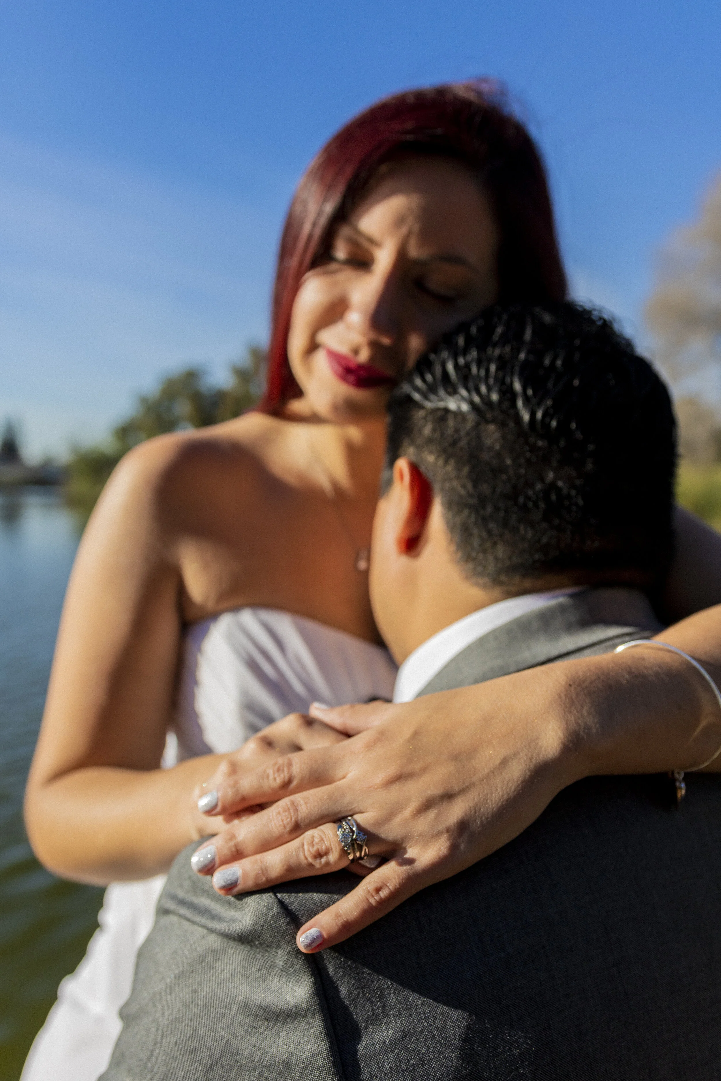 A woman with red hair embracing a man wearing a suit outdoors near a body of water on a sunny day.