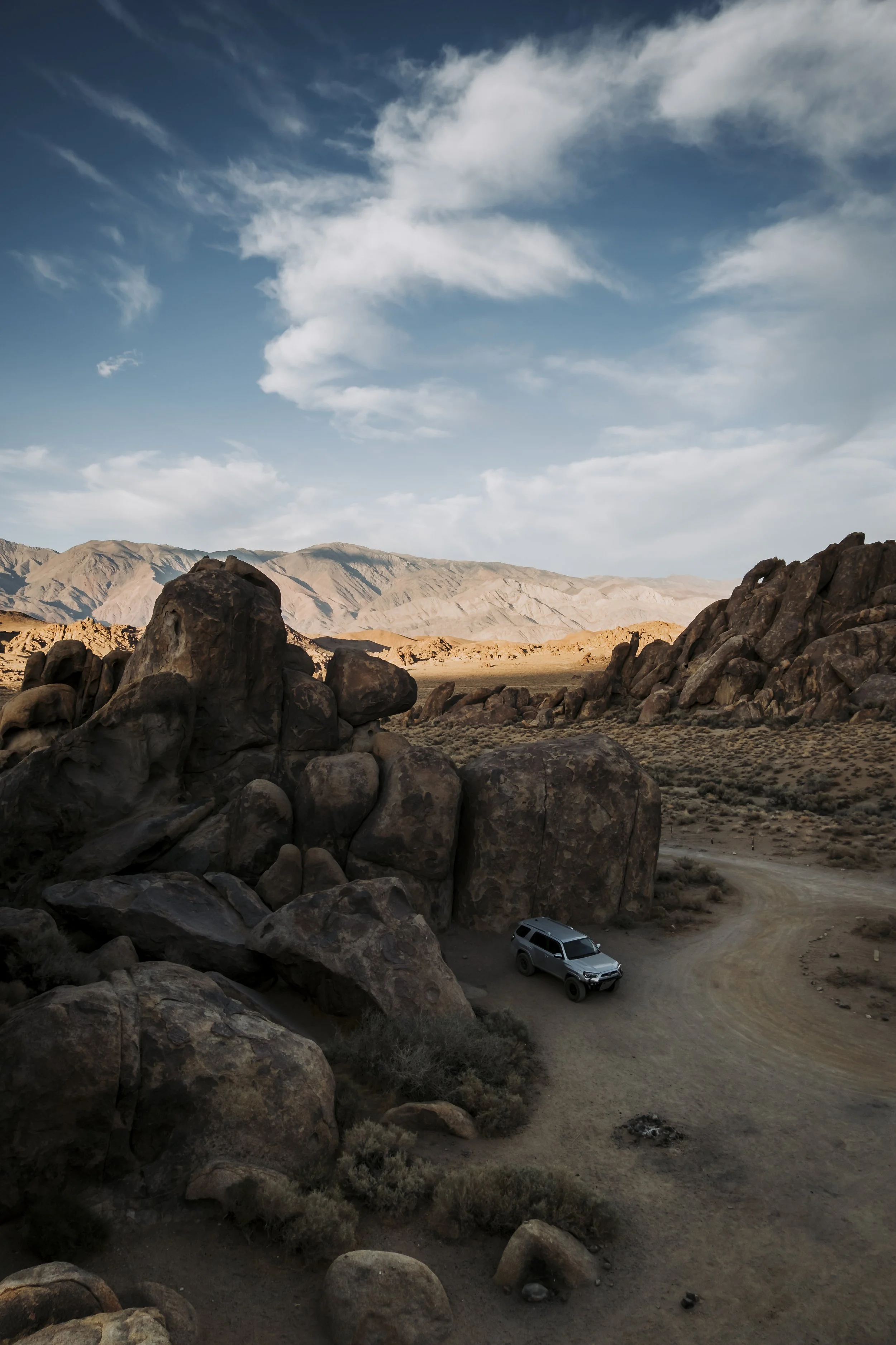 A silver SUV parked on a dirt road in a rocky, desert landscape with mountains in the background and a partly cloudy sky.