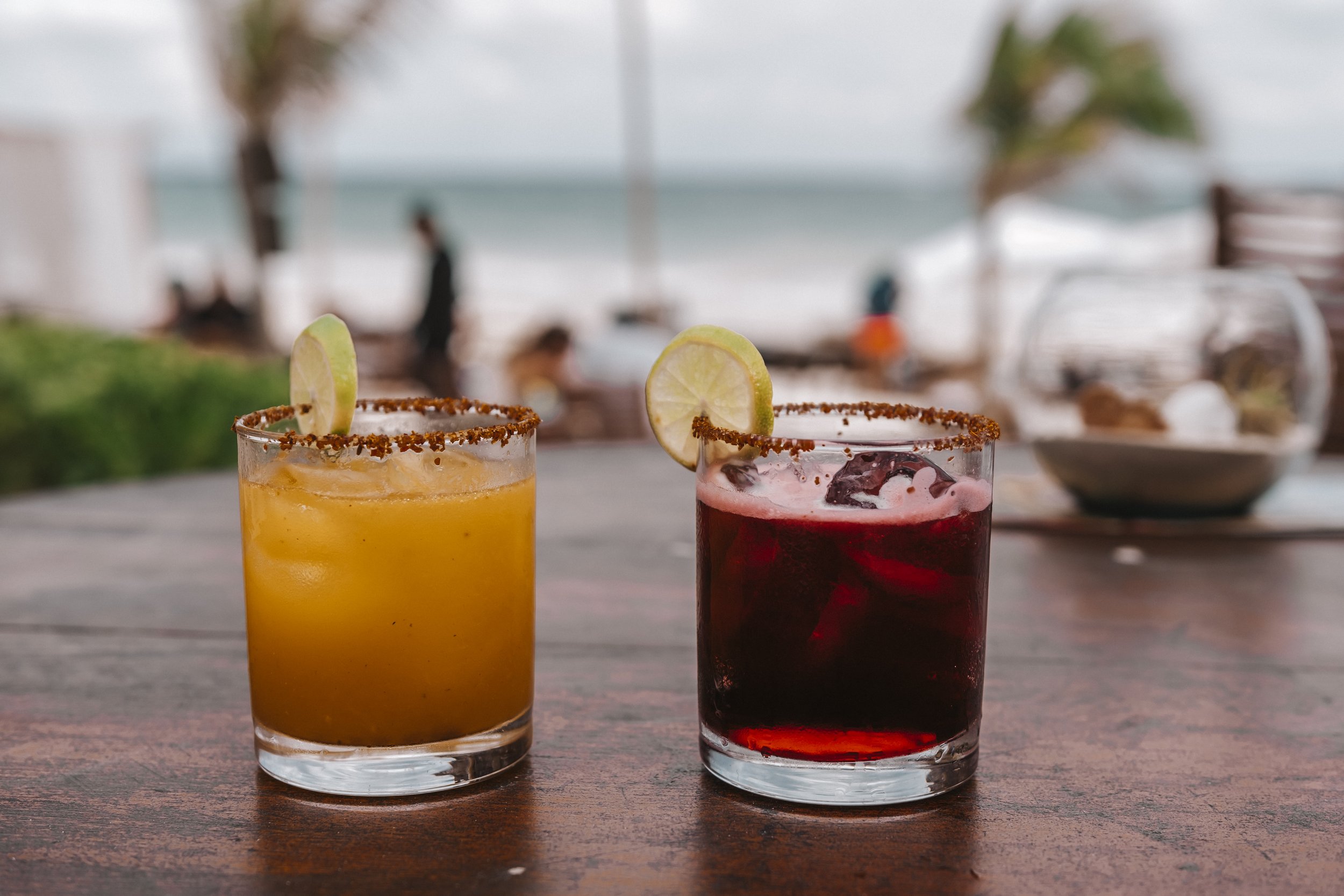 Two colorful cocktails on a wooden table with a beach and ocean in the background, garnished with lime slices and rimmed with chili powder.