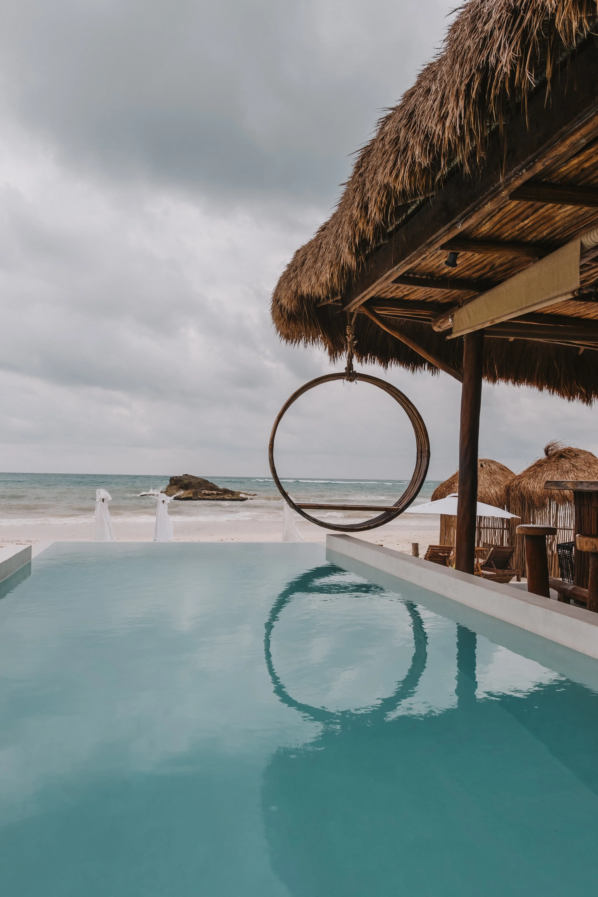 A tropical beach with a swimming pool, thatched roof huts, and a hanging circular swing, under a cloudy sky.