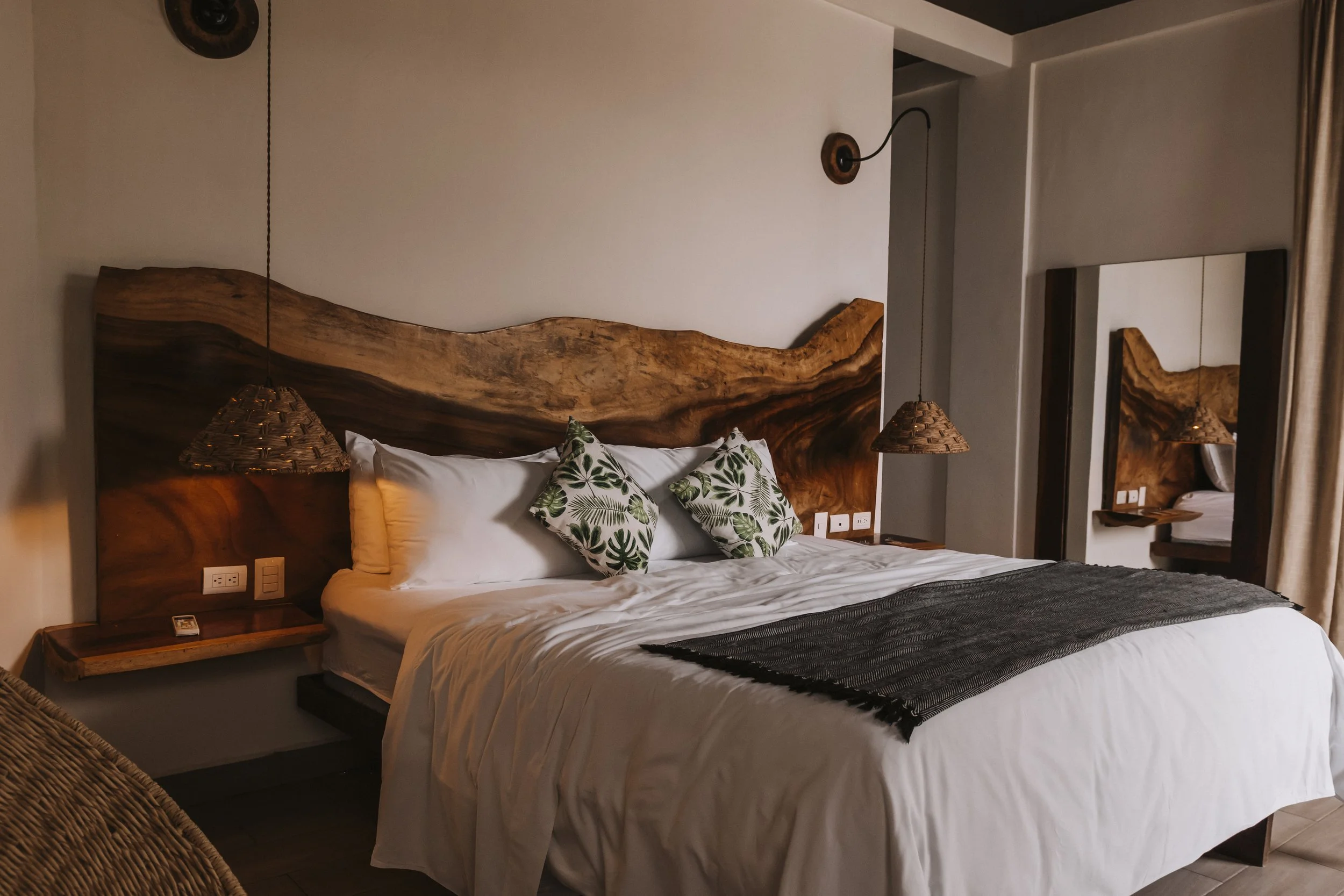 A hotel room with a bed, two green and white patterned pillows, a dark runner on the white bedspread, a wooden headboard, hanging woven lamps, a mirror, and a side table.