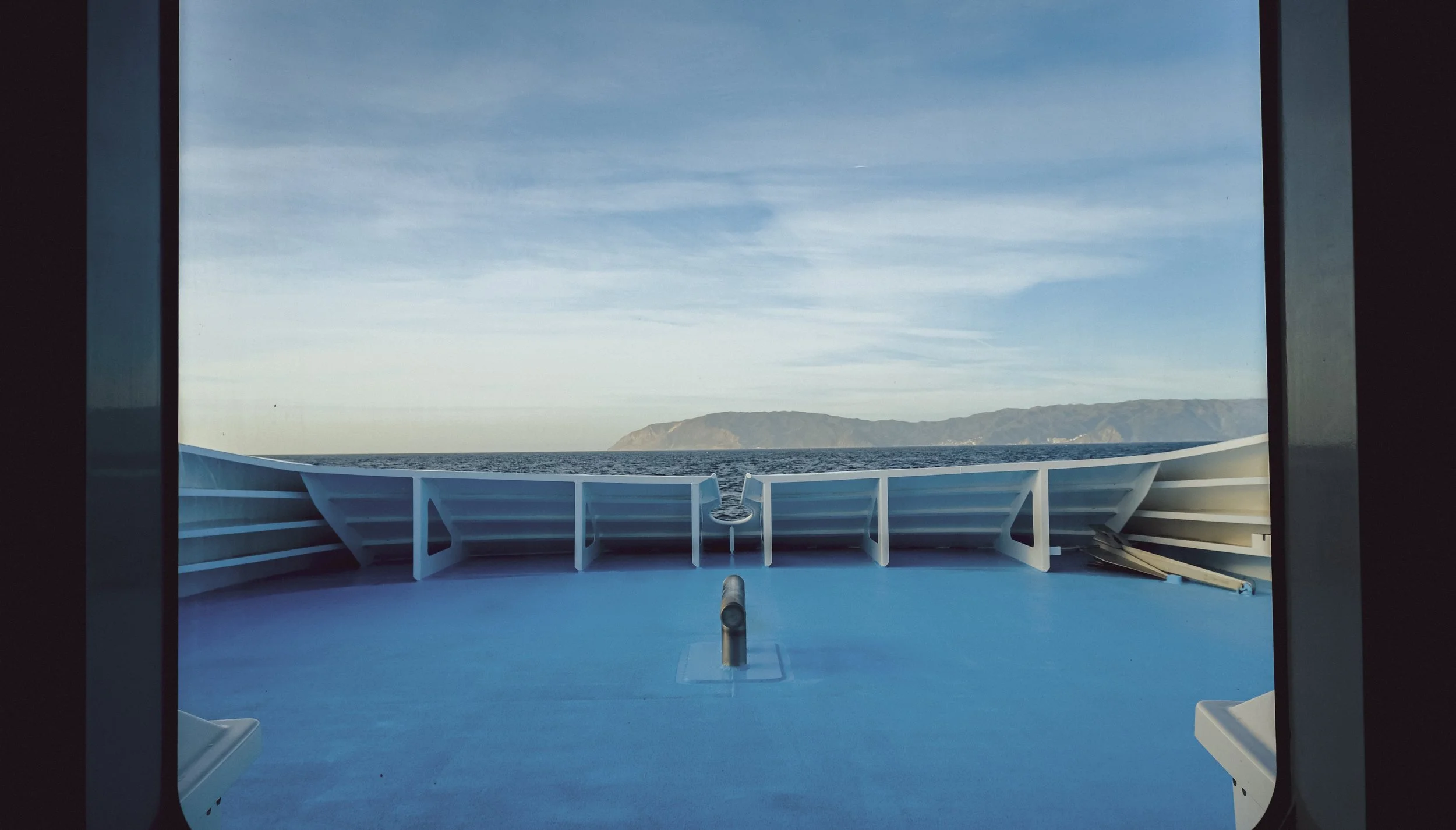 View from a ship's deck with a bright blue floor, white railings, and an ocean with distant hills under a partly cloudy sky.