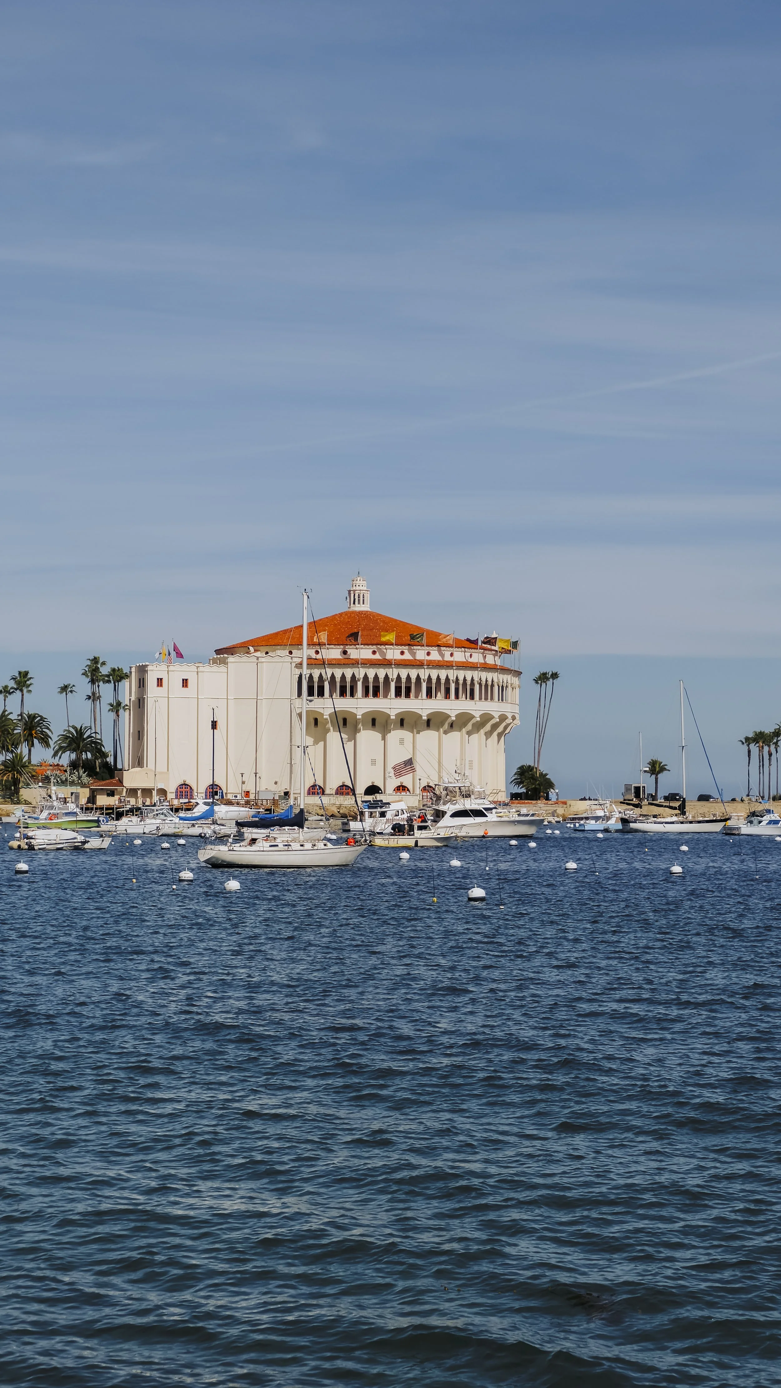 A waterfront scene with a large white circular building with a red-tiled roof, anchored boats in the water, and palm trees in the background under a clear blue sky.