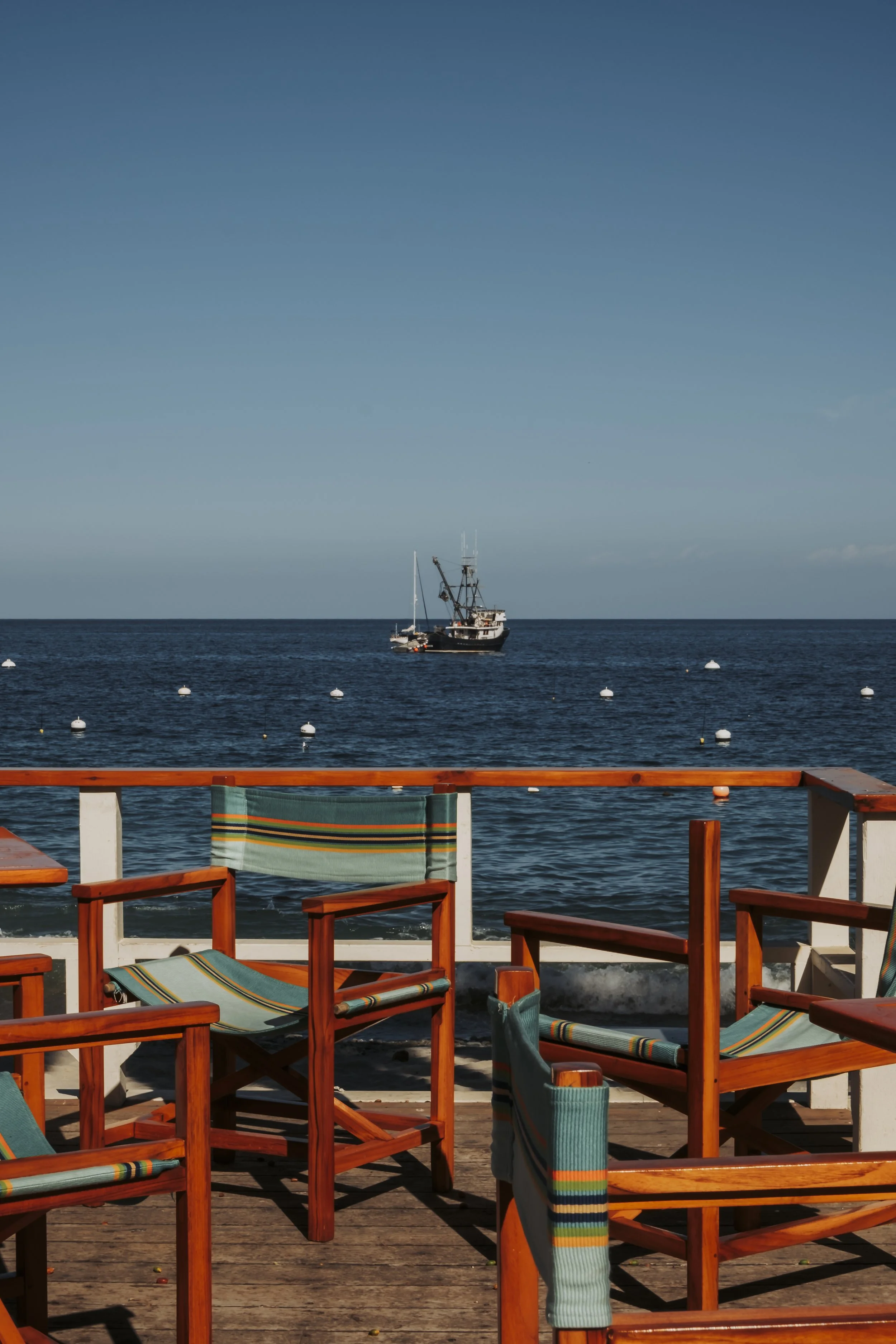 View of a wooden deck with striped chairs overlooking the ocean, with a boat anchored in the water and a clear blue sky.