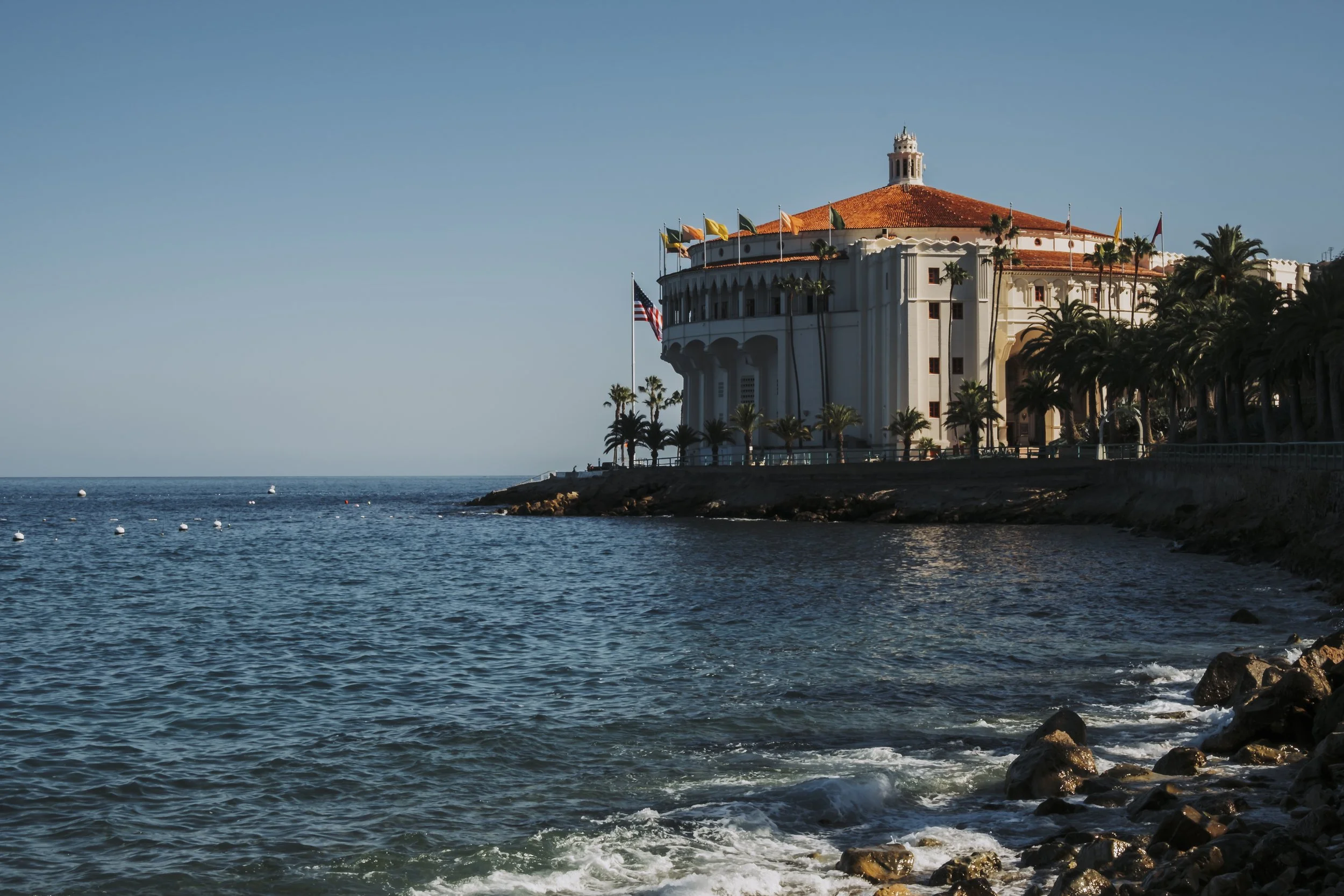 A large white building with a red-tiled roof near the ocean, surrounded by palm trees, with an American flag flying on a flagpole, set against a clear blue sky.