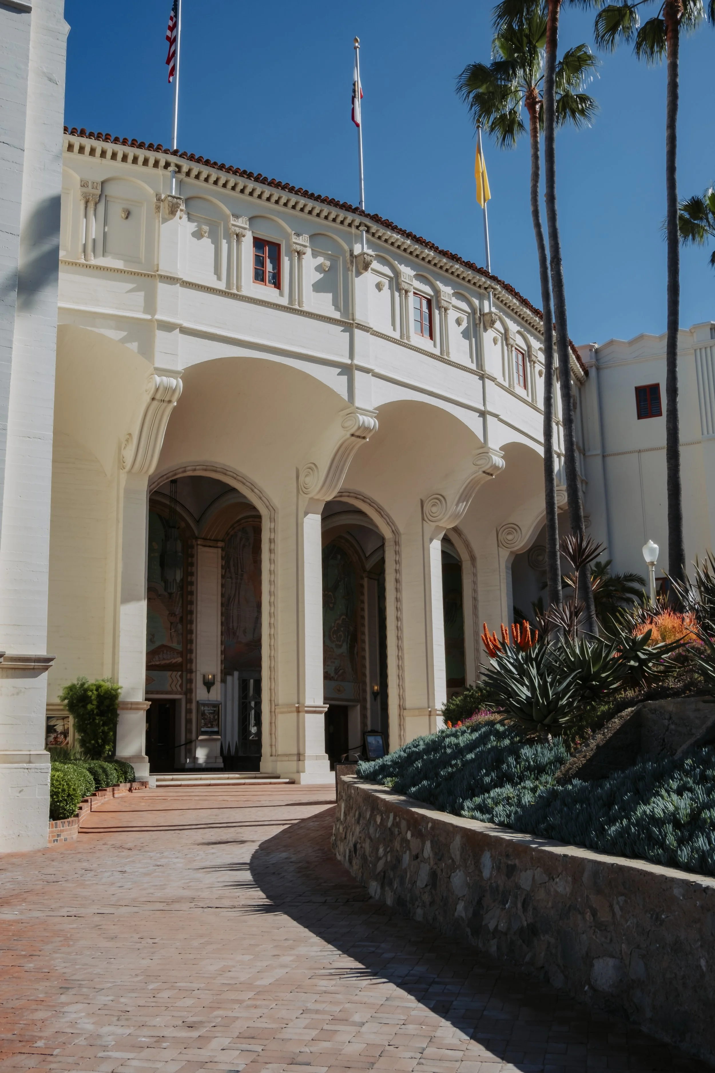 Facade of a historic building with arched entrance, white walls, decorative architectural details, flags, and palm trees against a clear blue sky.