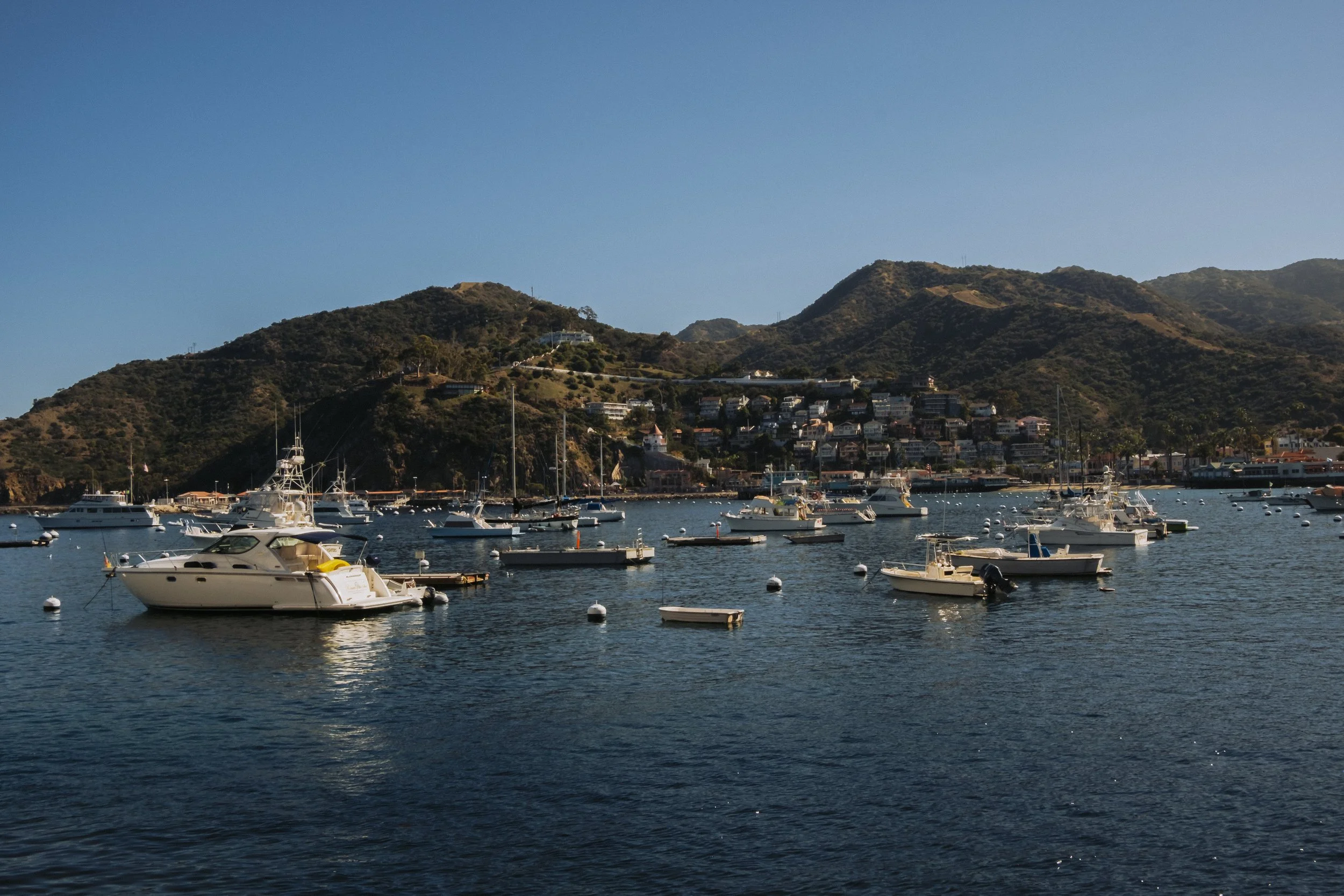 Many boats and yachts anchored in a harbor with a hillside town and mountains in the background during daytime.