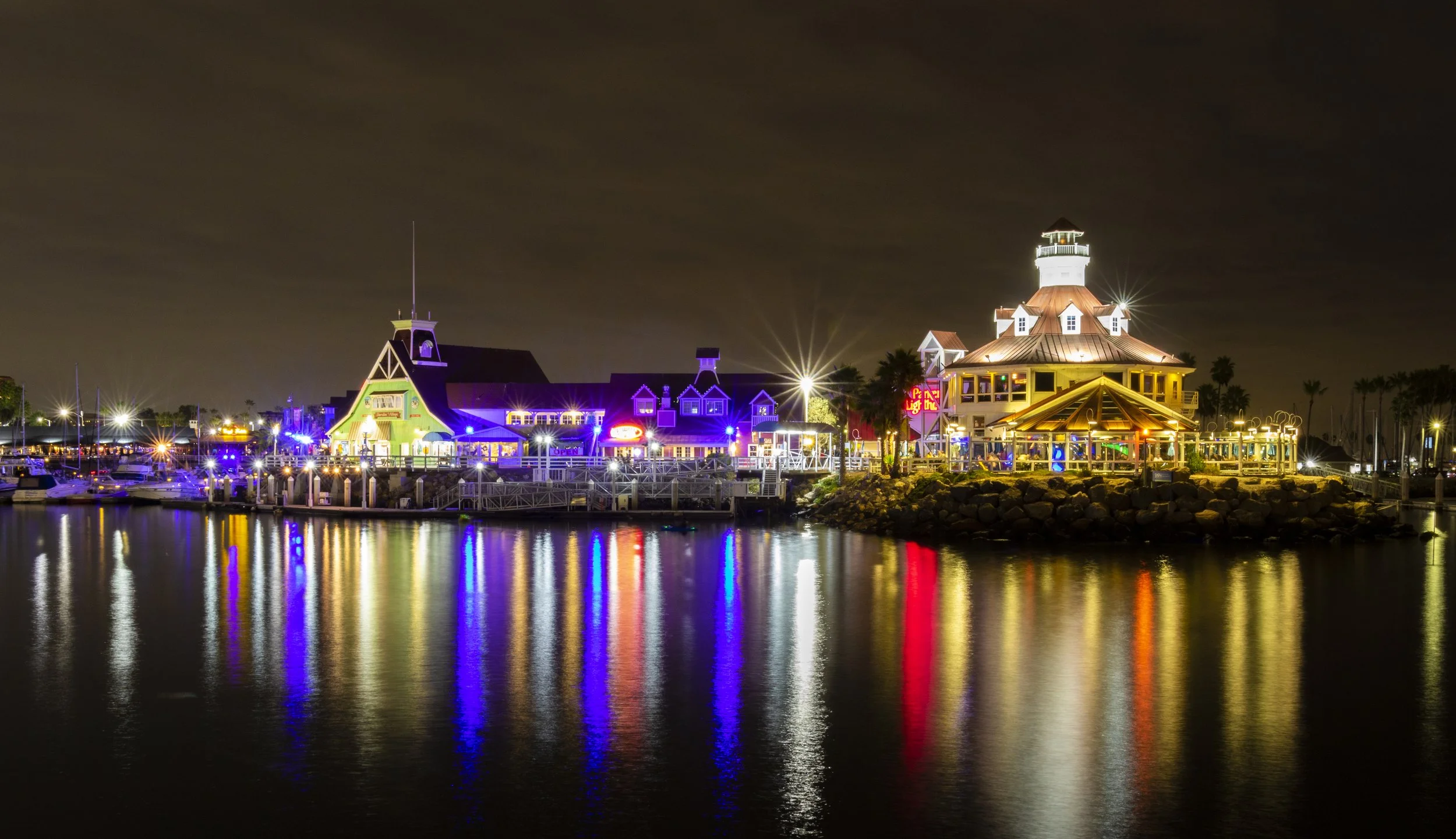 Night view of colorful illuminated buildings and boats reflected on calm water, with a lighthouse-like structure on a rocky pier.