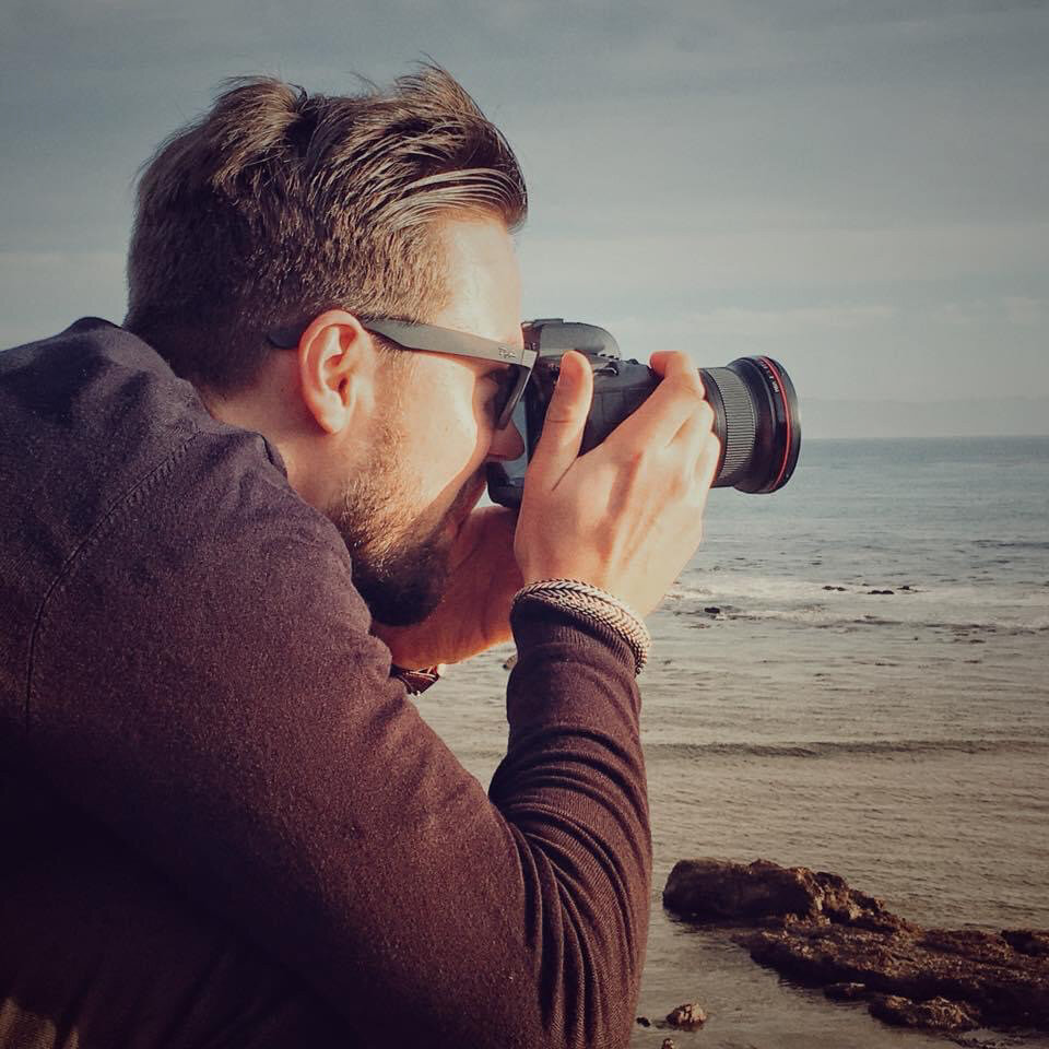 A man with sunglasses and a beard is taking a photo with a camera by the seaside, with rocks and the ocean in the background.