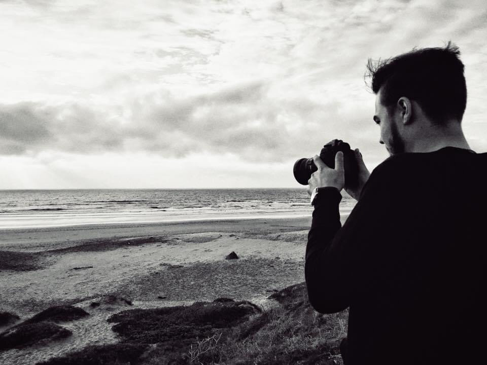 A man taking a photograph on the beach, with the ocean and cloudy sky in the background, in black and white.