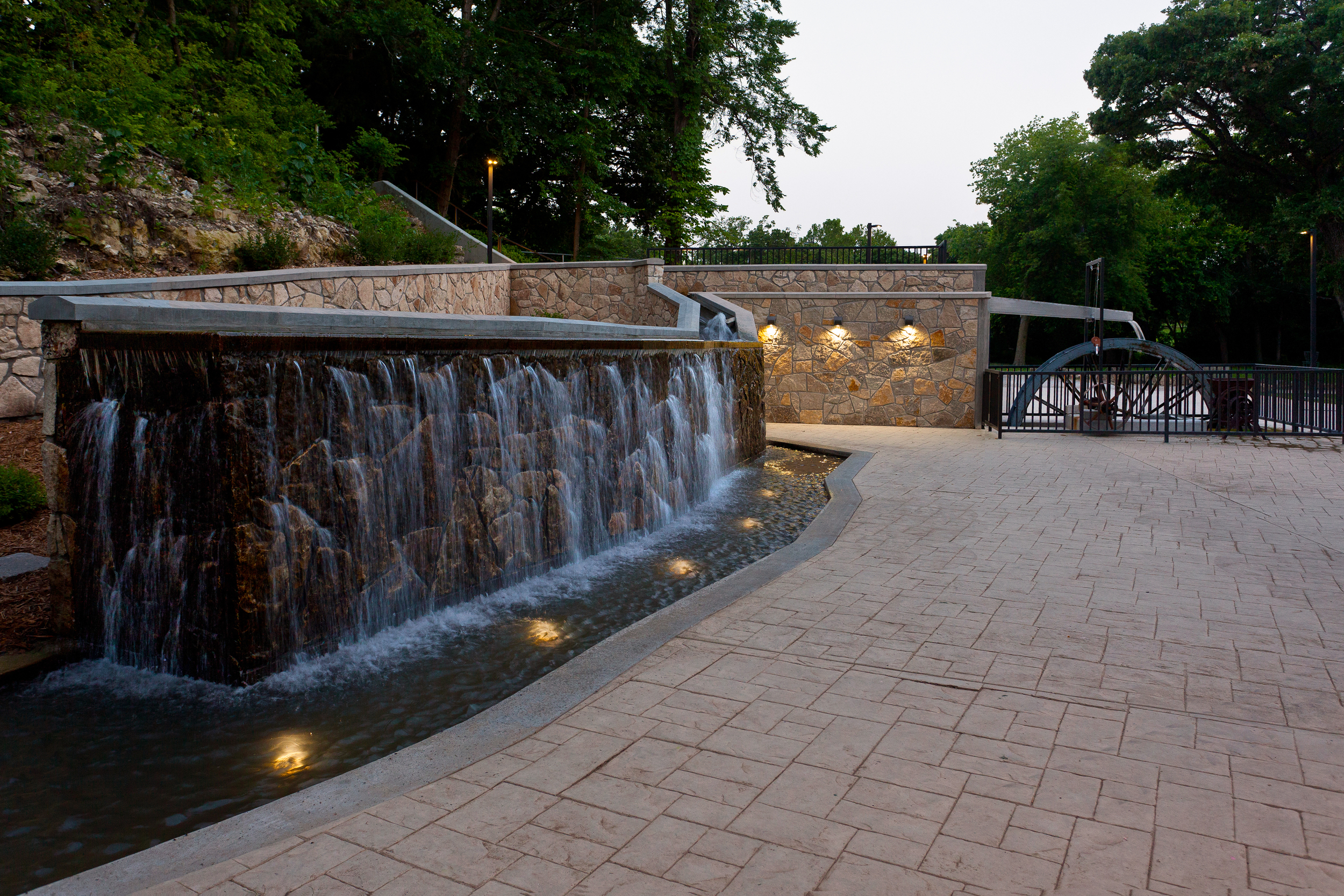 The spring emerges from the hillside and flows through two flumes; the upper flume turns the new water wheel as shown in the background; the lower flume, in the foreground, flows through an open channel before cascading across the face of a stone wall. The lower flume provides a place for campers and retreaters to interact with the water as it flows over the stone wall.
