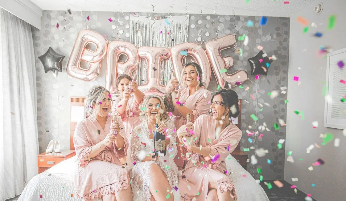 Bride with her bridesmaids popping confetti and champagne while wearing robes on her wedding day. Fun wedding photo that is light and airy. 