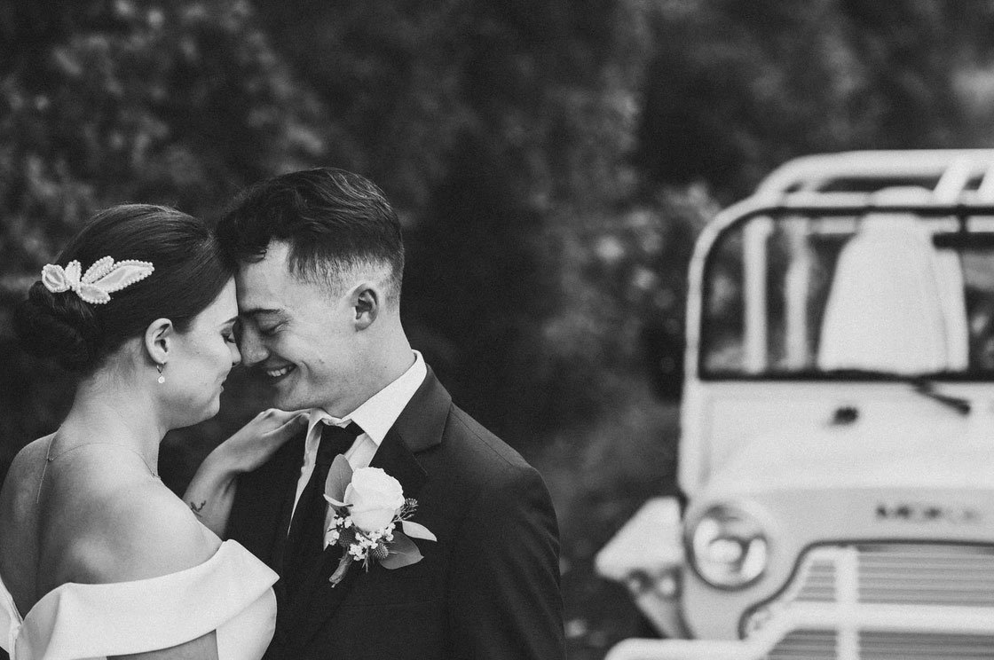 B&W wedding photo of a bride & groom touching foreheads with a vintage car in the background at Bear Brook Valley wedding venue. 