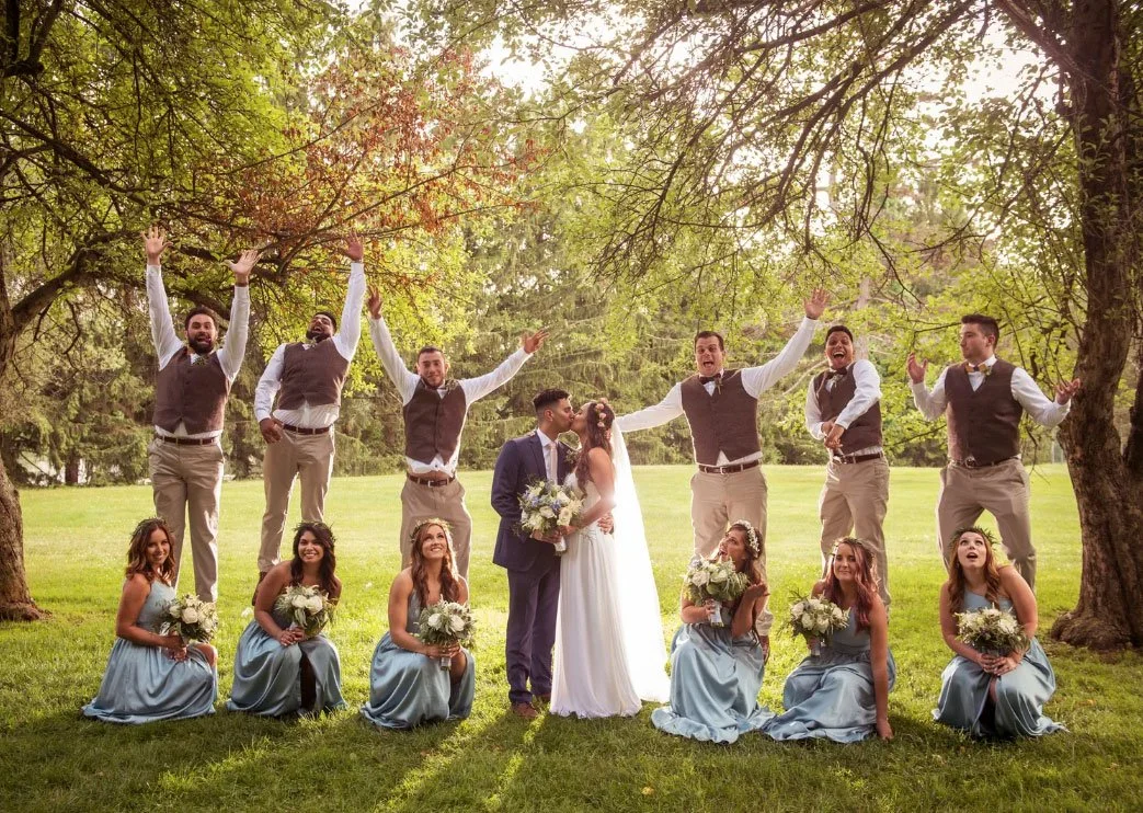 a Perona Farms wedding couple shares a kiss in the woods while the bridal party looks on and is cheering. 
