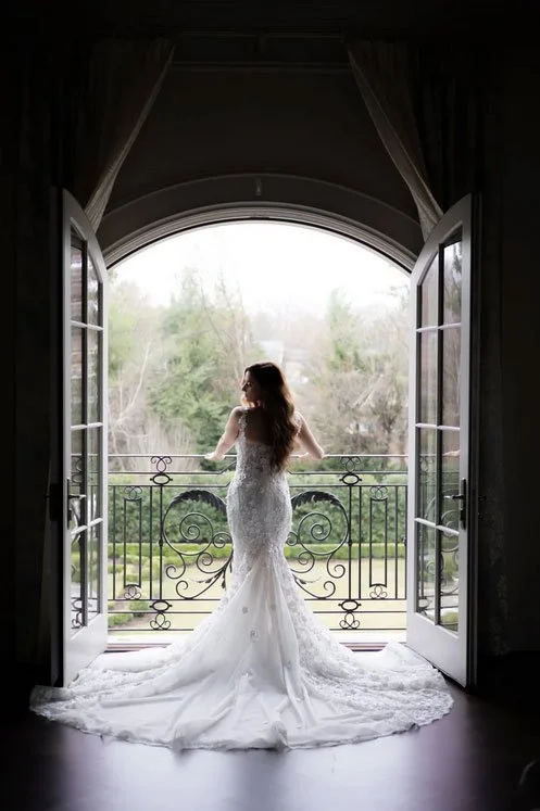 Park Chateau bride looks out at gardens of her wedding venue from the bridal suite. Her dress is all fanned out, and the wedding photo is documentary style with natural lighting & color. 