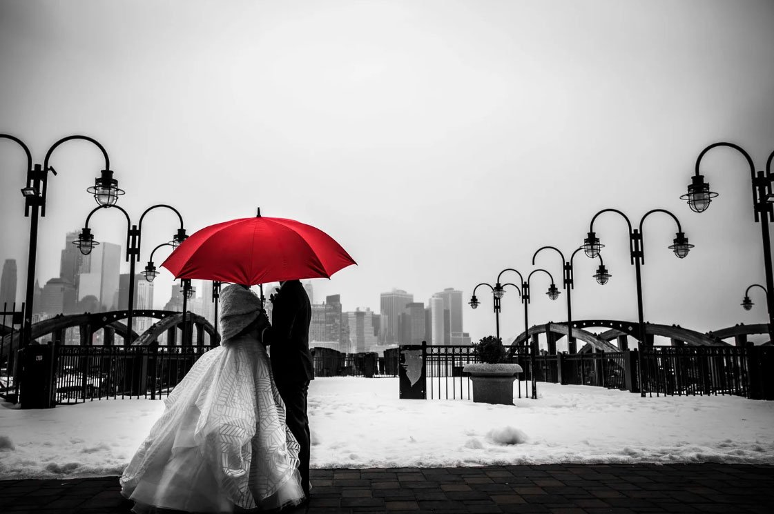 Gabrielle Delia a New Jersey Wedding Photographer captures black and white photo of the bride & groom as they walk on the pier in Jersey City with a red umbrella. 