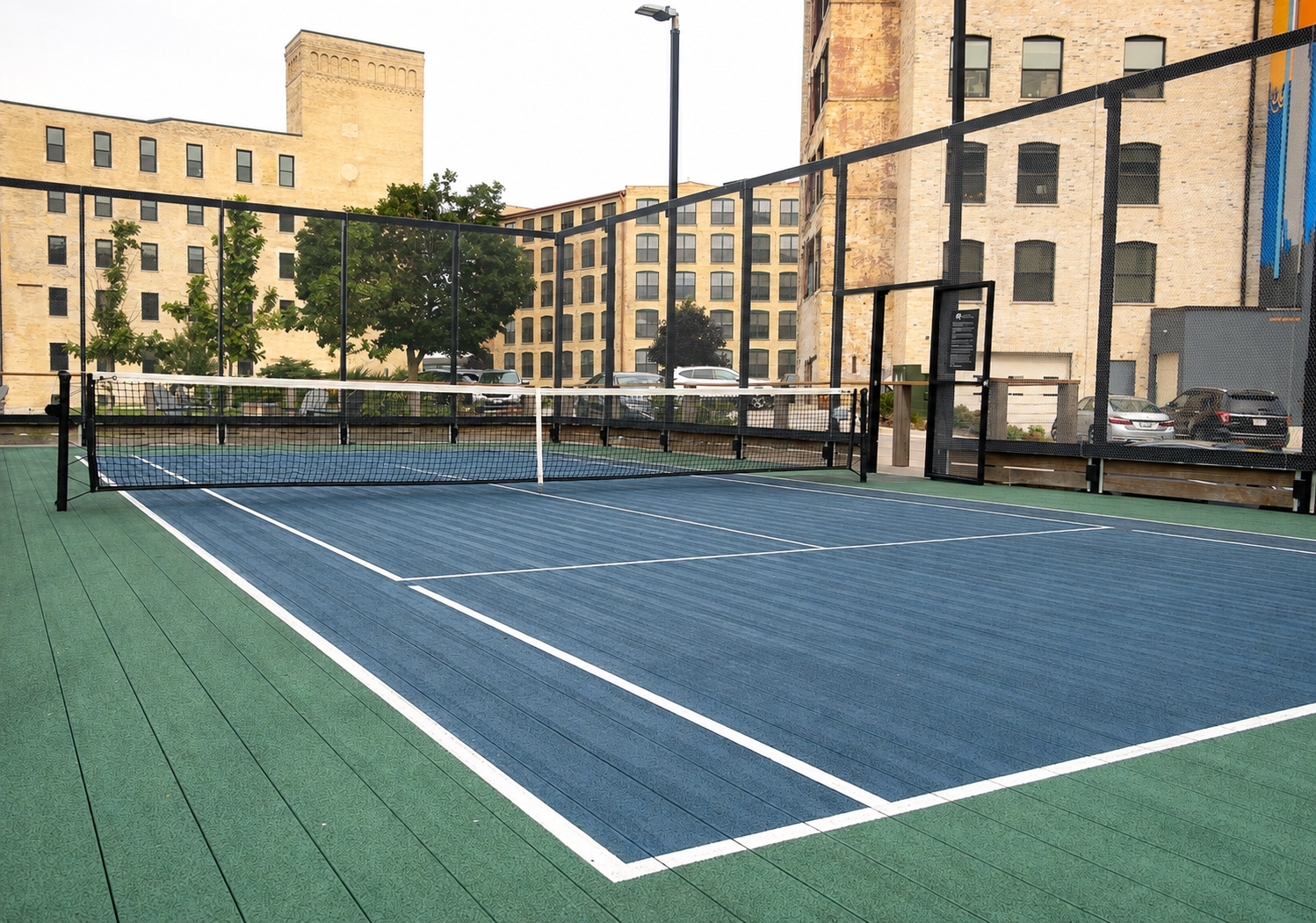 An empty outdoor pickleball court with blue playing surface and white boundary lines, surrounded by a black fence and located in an urban area with beige brick apartment buildings and parked cars in the background.
