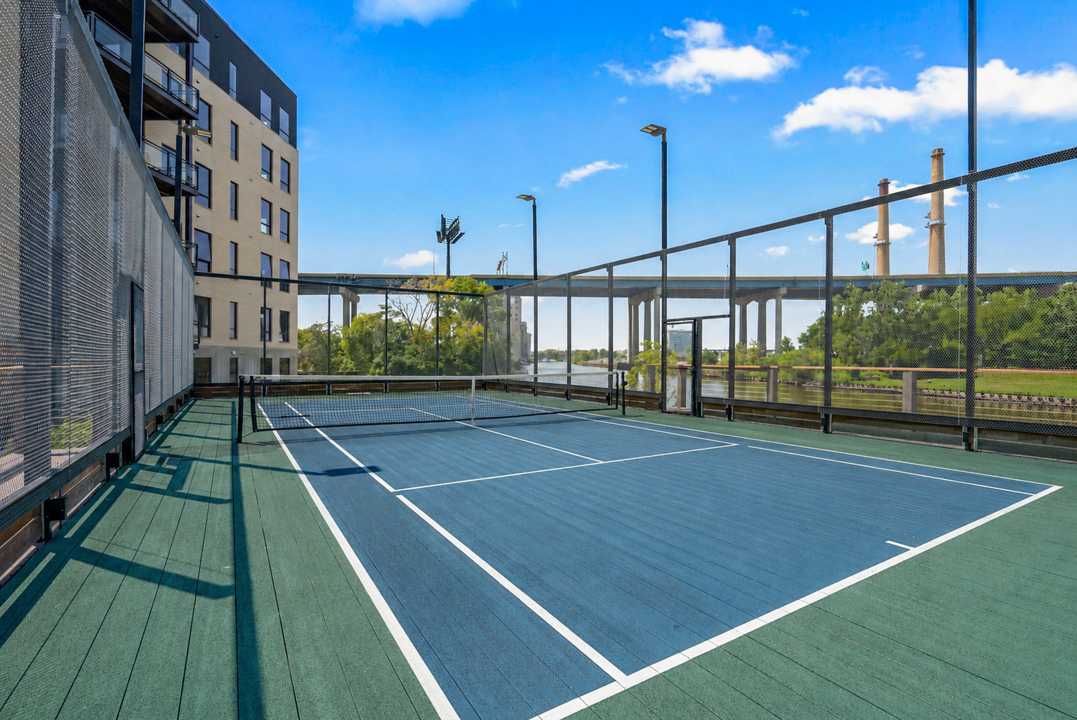 Outdoor rooftop tennis court with blue and green surface, surrounded by a safety net and fencing, situated next to a modern multi-story building under a clear blue sky with trees and industrial chimneys in the background.