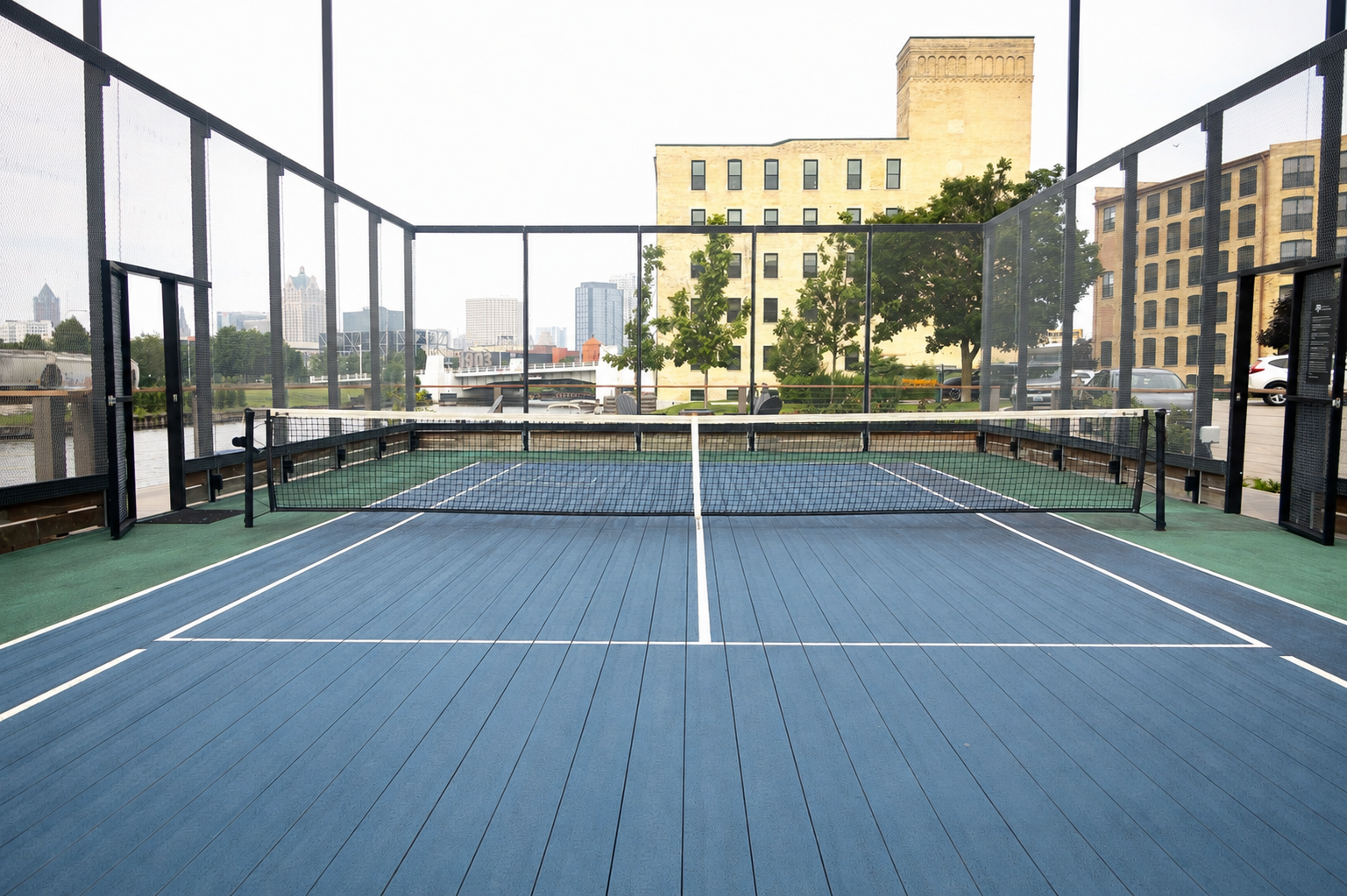 Outdoor rooftop pickleball court with a net, surrounded by a safety netting, with city buildings and parked cars in the background.