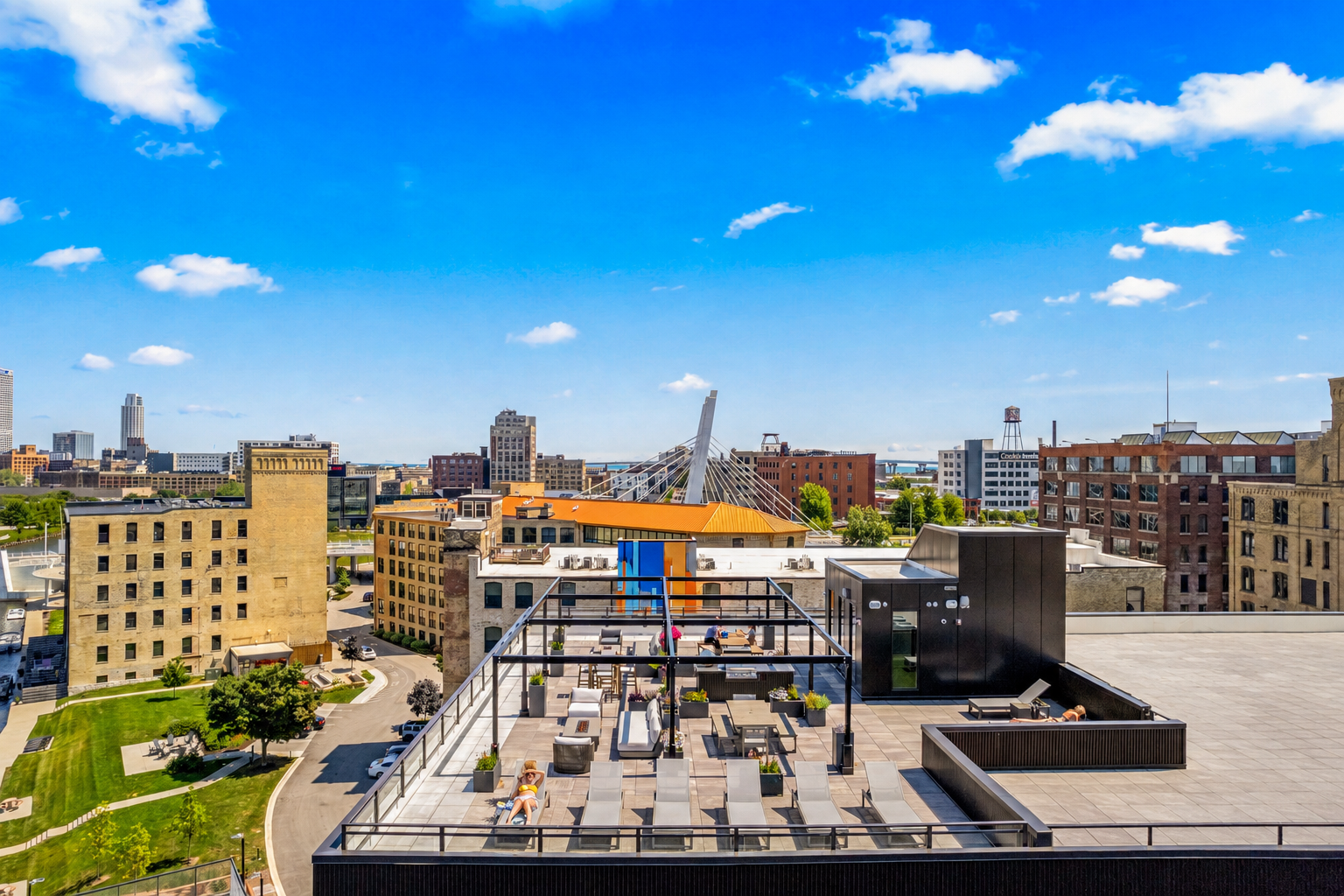 City skyline with modern buildings, a bridge, and a rooftop terrace with lounge chairs, tables, and people relaxing under a blue sky with scattered clouds.