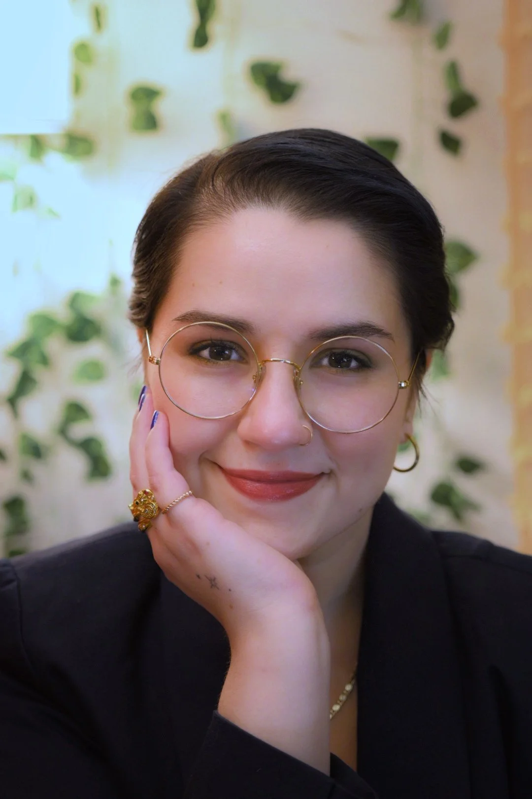 A portrait of a woman with short dark hair, brown eyes, and round gold glasses smiles calmly ahead while resting her chin in her hand.