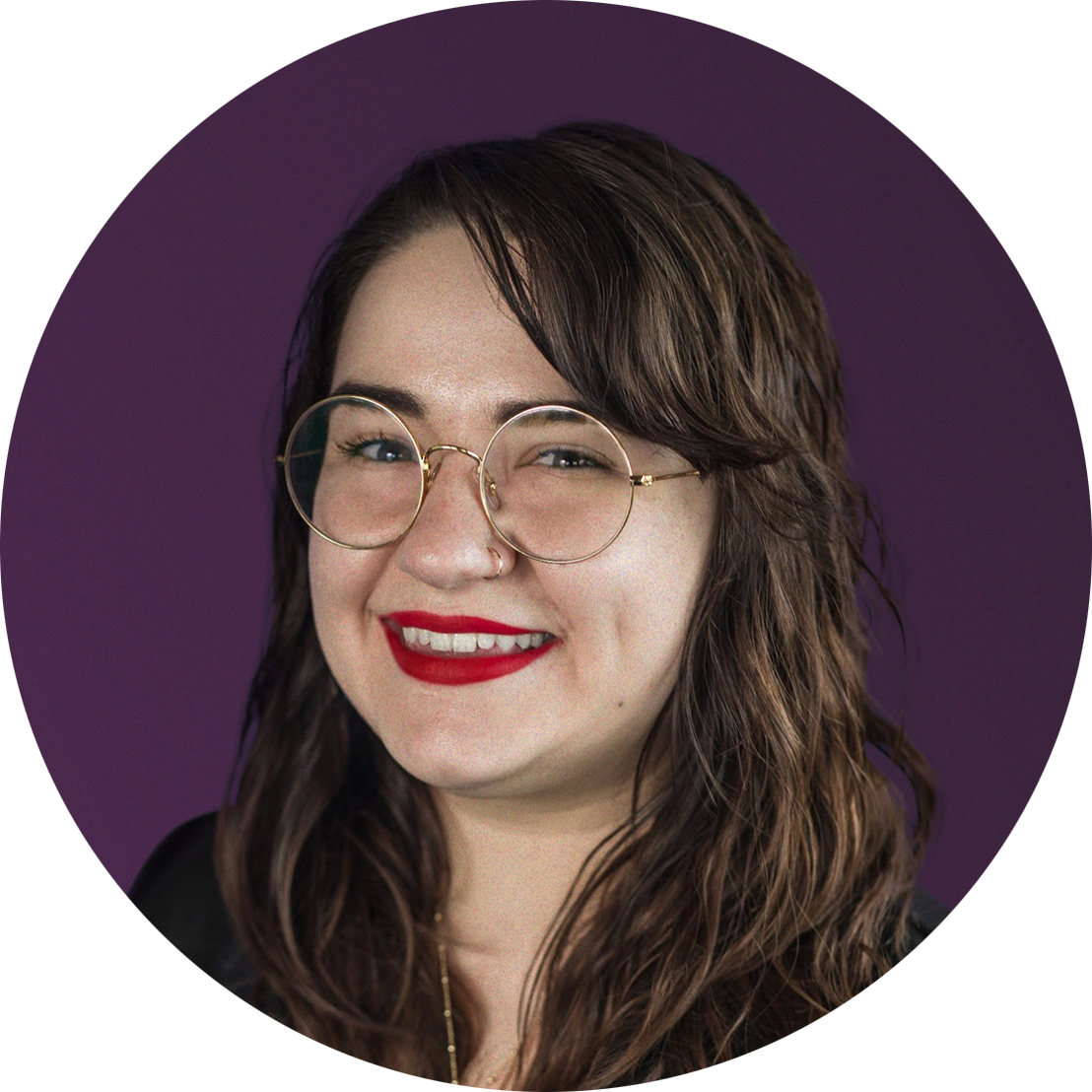A young woman with long, wavy brown hair, wearing round glasses, red lipstick, and a nose ring, smiling against a purple background.