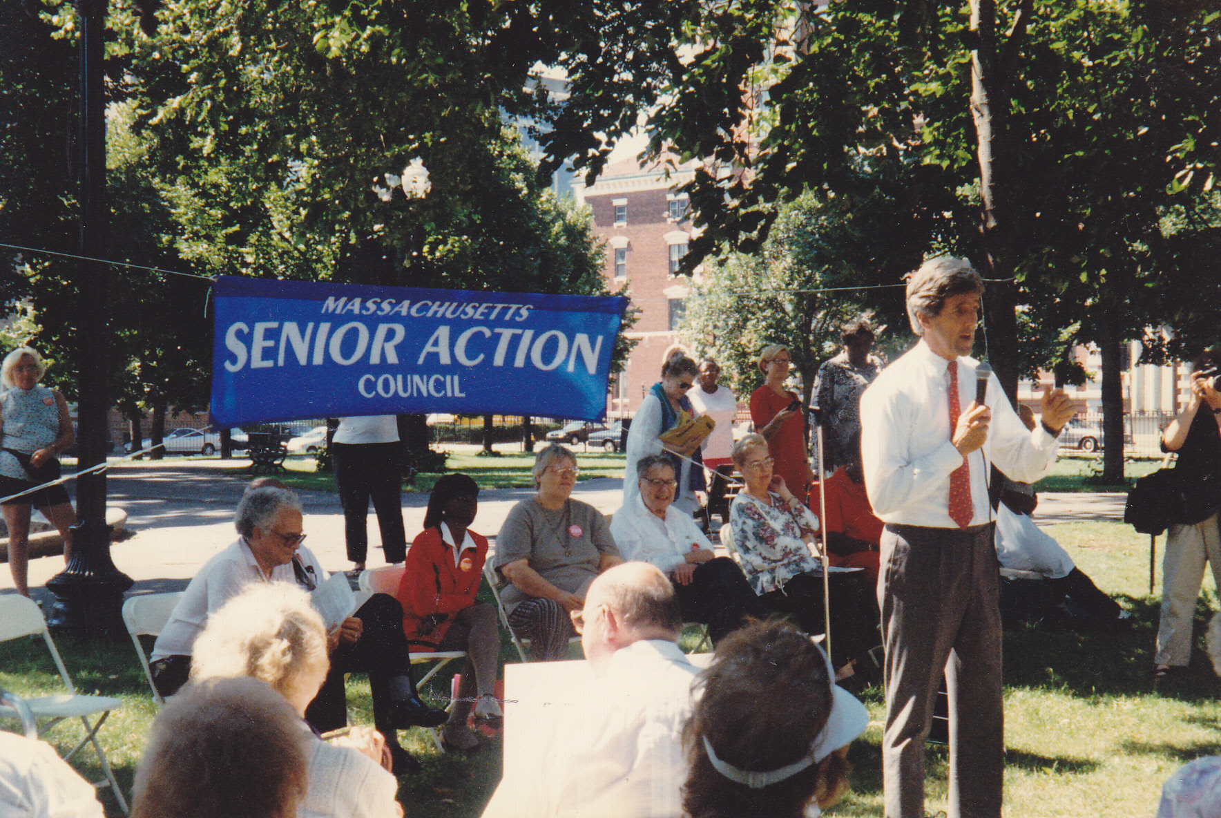 Man with a mic in a field with people gathered around and a Massachusetts Senior Action banner in the background