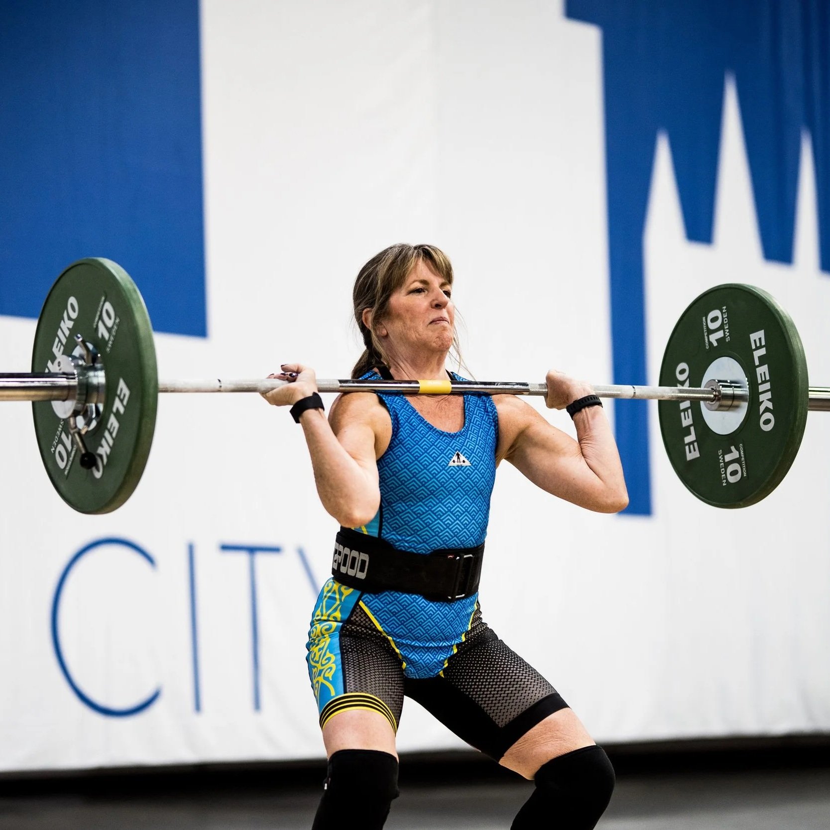 A older woman performing a clean and jerk with a barbell at a weightlifting competition.