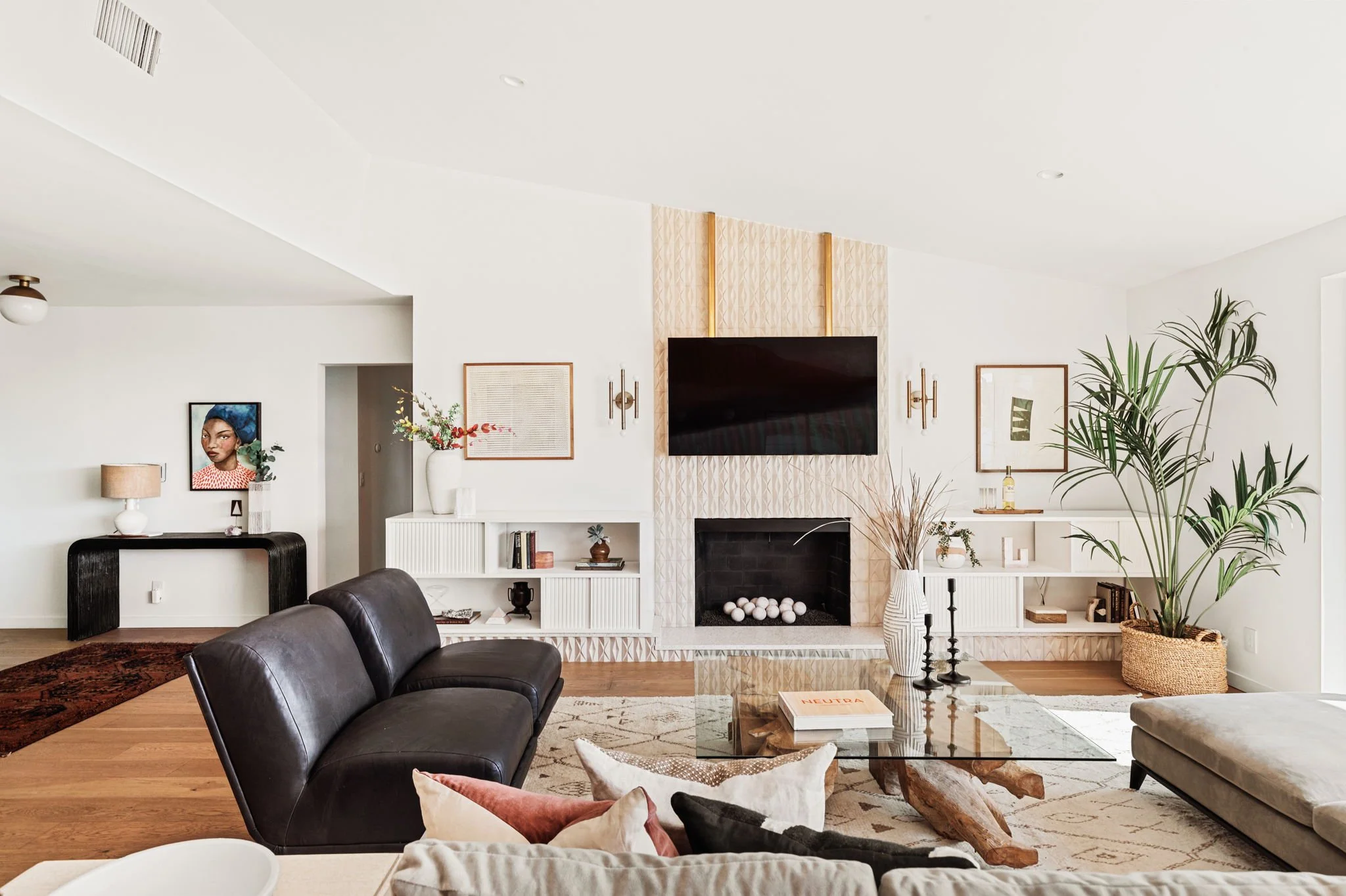  The contemporary stone fireplace in this mid-century living room is accented by two black leather club chairs and a large tree trunk and glass coffee table. Two pieces of neutral art hang on either side of the mounted TV above the fireplace, and the
