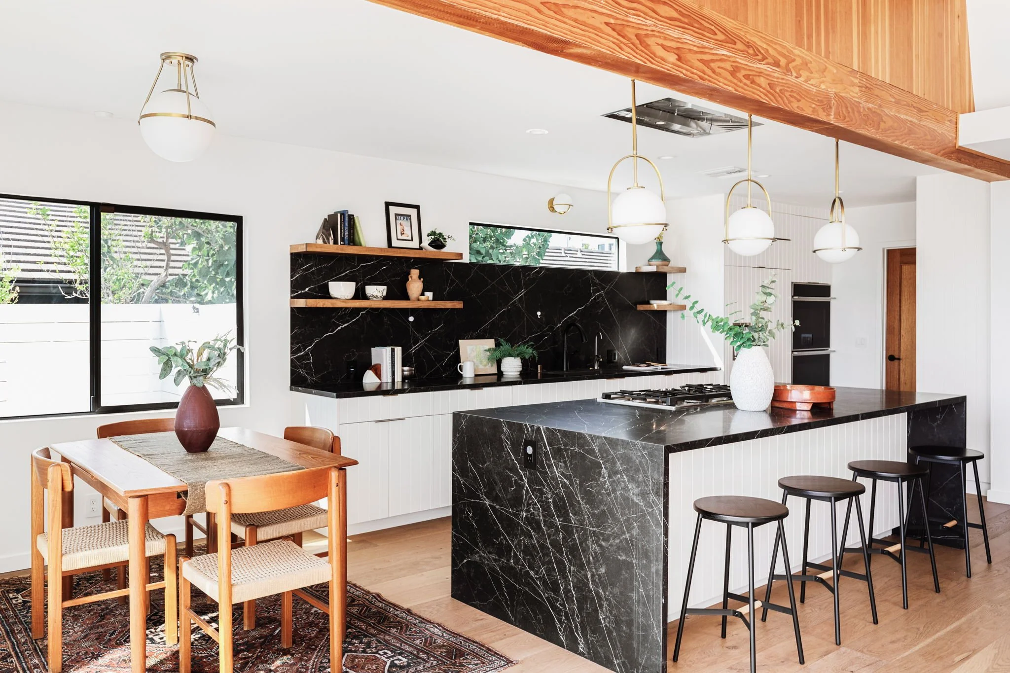  Contemporary kitchen and dining area featuring a large island with black marble countertops and black bar stools. Dining space has a wooden table and chairs set on a patterned rug. 