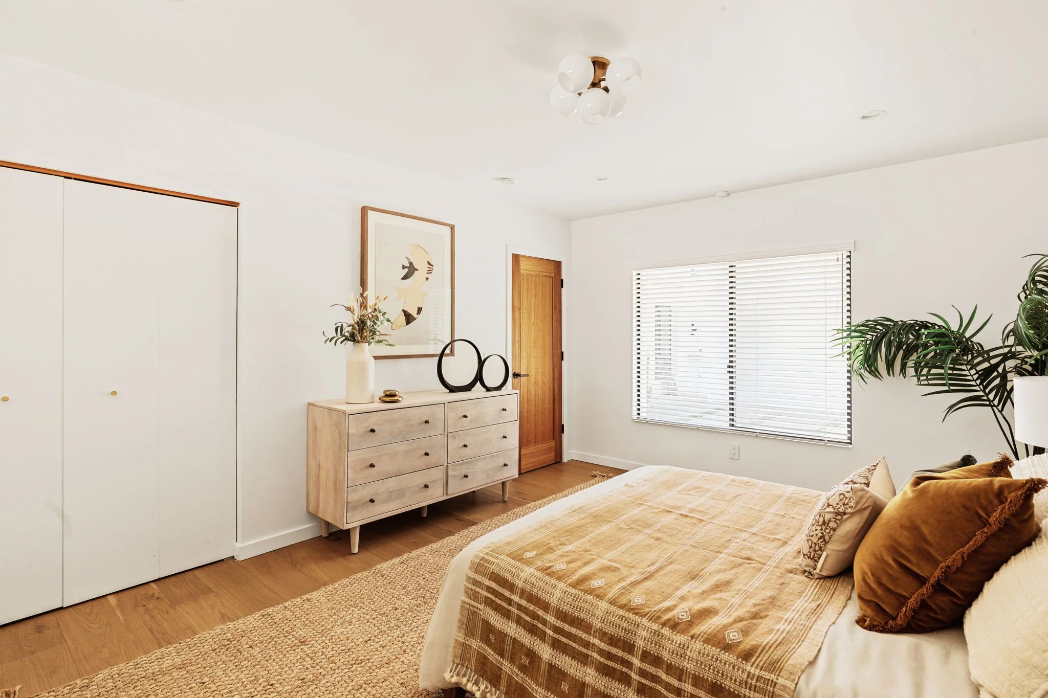  A neutral bedroom with a light wood dresser. Mustard pillows and a patterned throw blanket on the bed, and a jute rug. 
