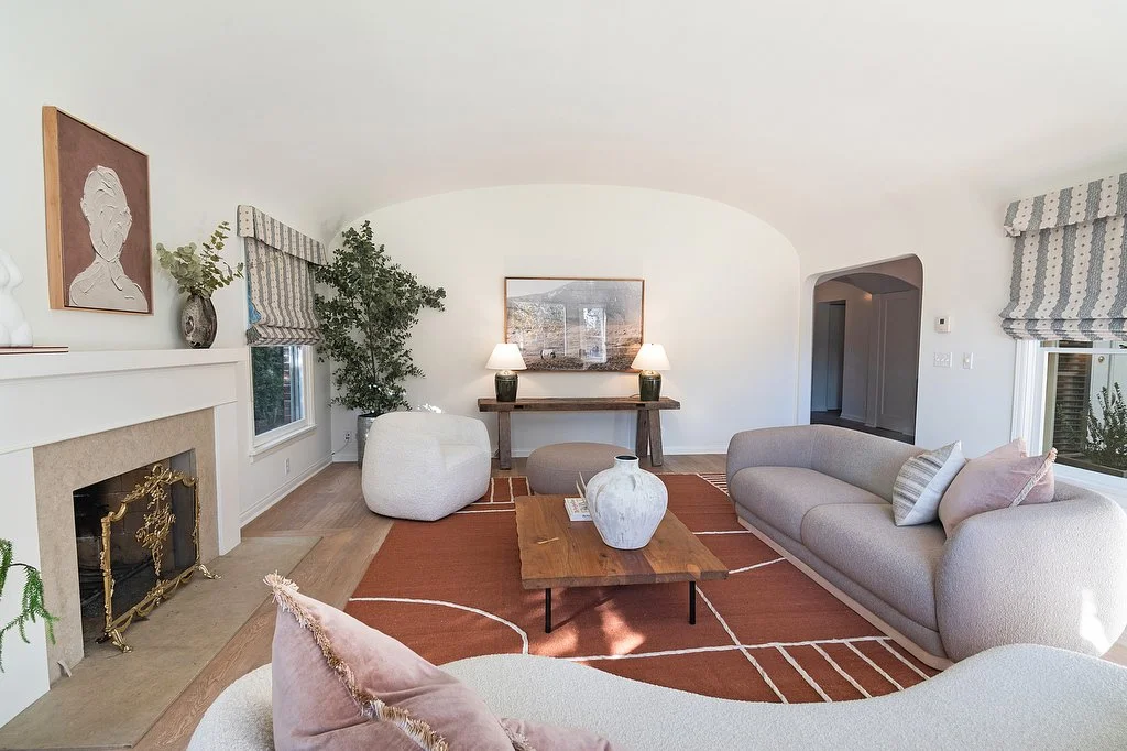  Curved furniture in neutral tones, centered around a wooden coffee table on a rust-colored geometric rug. Two table lamps on a wooden console. 
