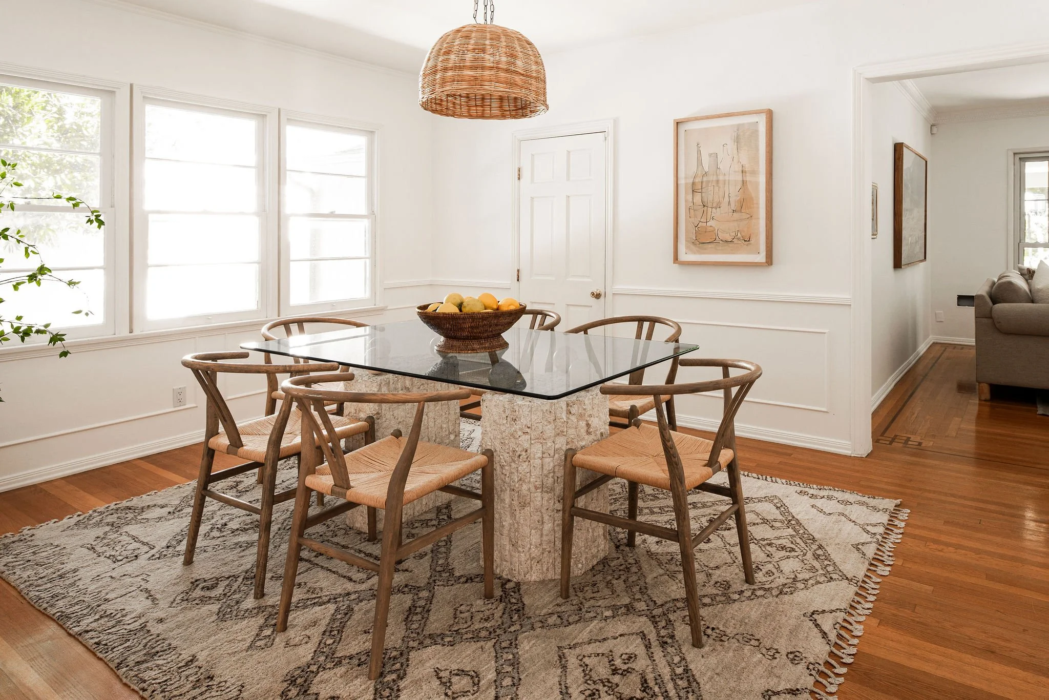  Modern glass-top table with a textured stone base, surrounded by natural woven chairs. A wicker pendant light hangs above, and an abstract rust painting hangs in the back. 