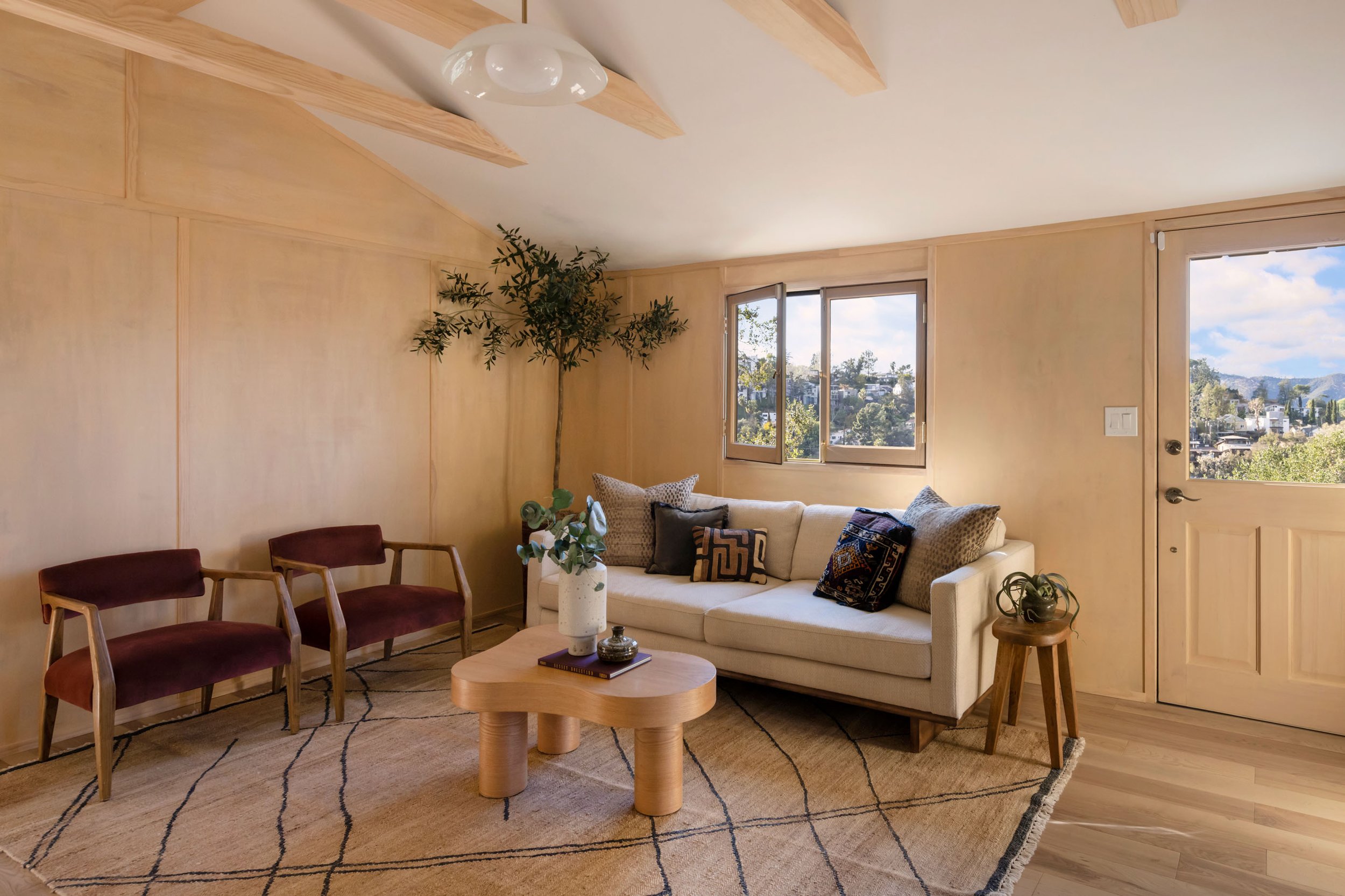  Living room surrounded in light wood paneling and beams. A neutral sofa accented with red and green textured throw pillows, sits on a jute rug with two wine colored upholstered chairs to the left. 
