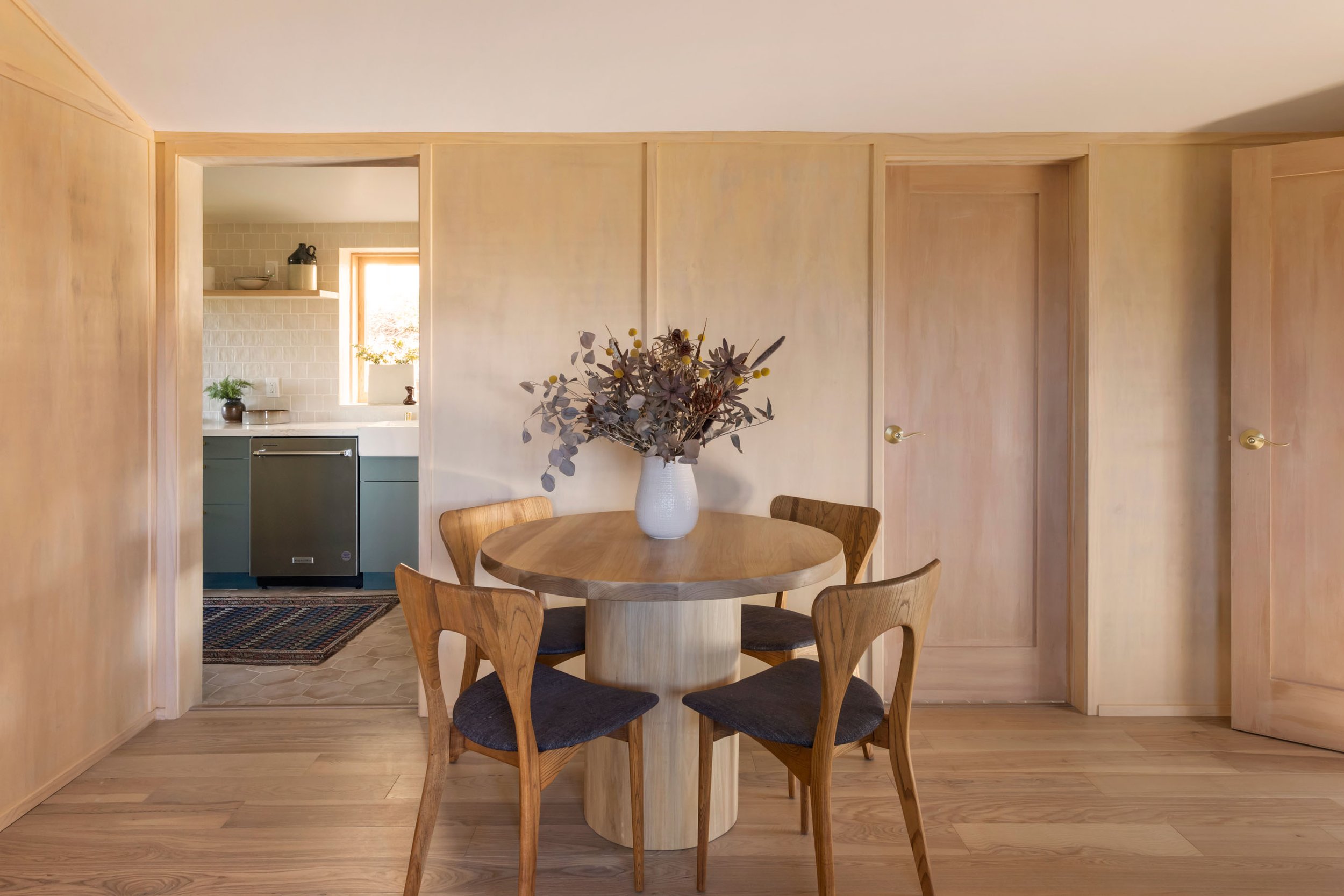  Light wood paneled dinging room with a round oak dining table surrounded by four wood and upholstered dining chairs. A large dried floral arrangement in a white ceramic vase sits in the middle of the table. 