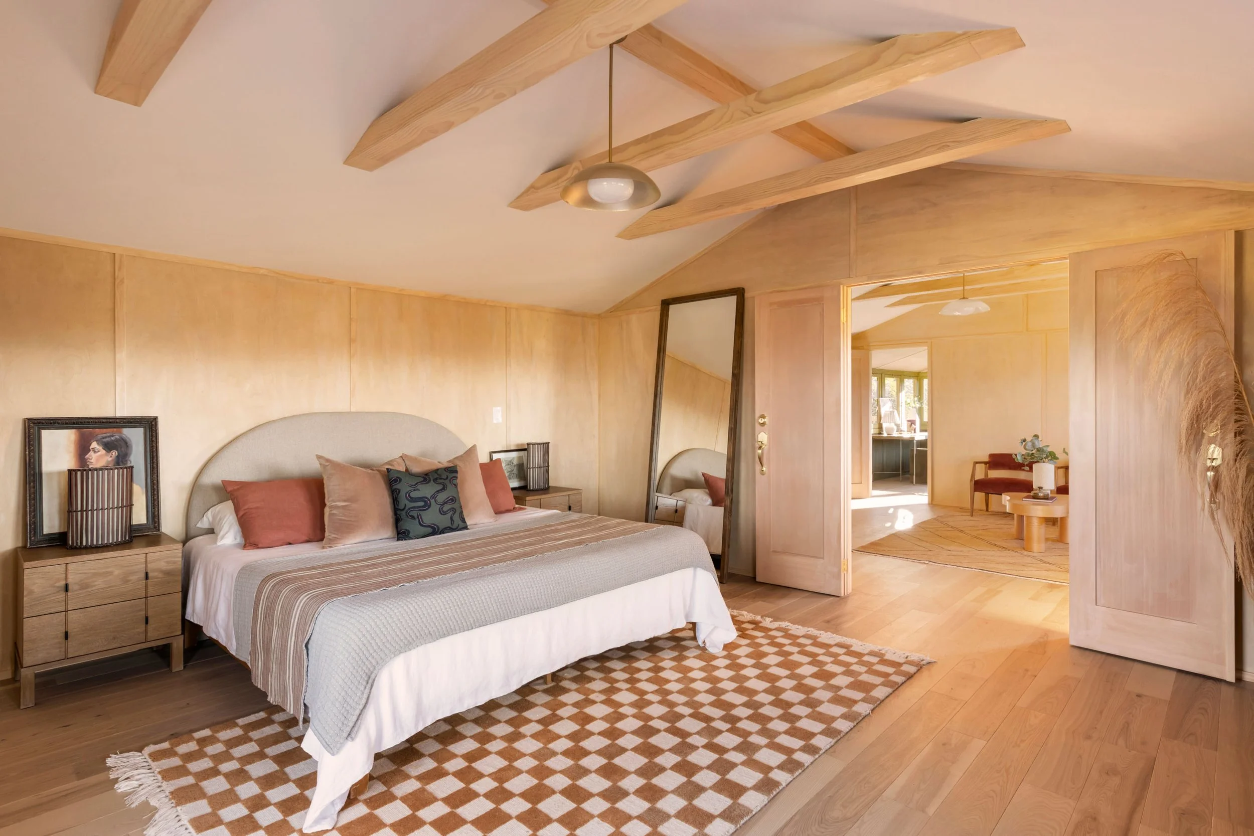  Primary bedroom with light wood paneled walls and beams, neutral bedding and arched headboard are accented with orange, pink, and green throw pillows. The bed sits on a white and orange checkered rug.  