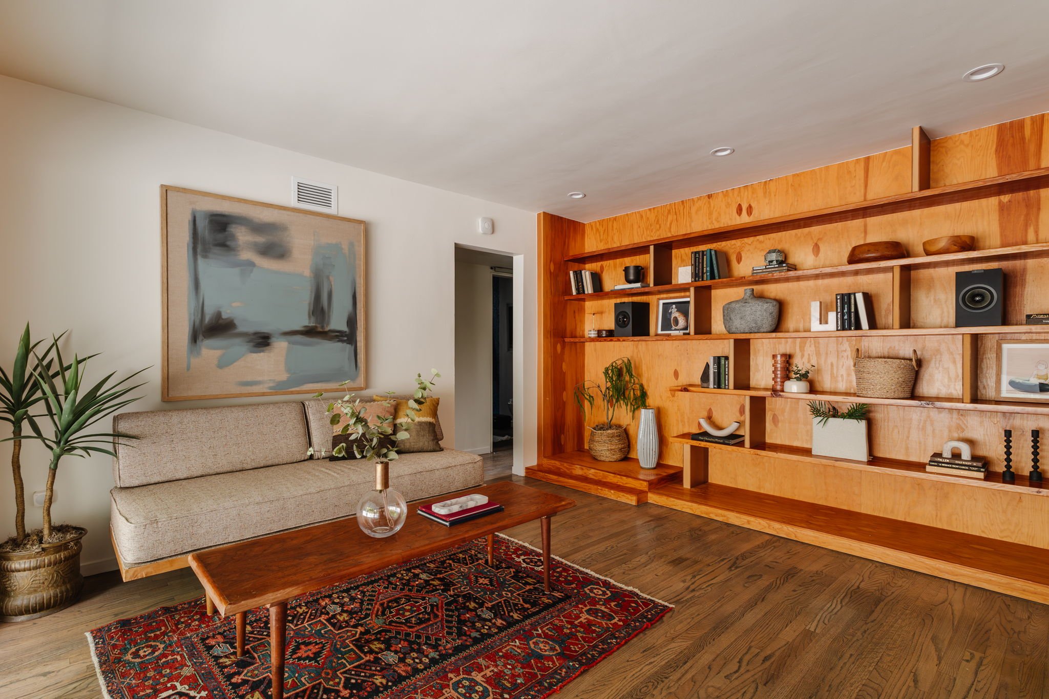  Wide shot of a solid wood built in bookshelf accessorized with books and found objects. A MCM daybed is to the left sitting on a vintage Persian rug, with a beige and blue abstract artwork hung above the daybed. 