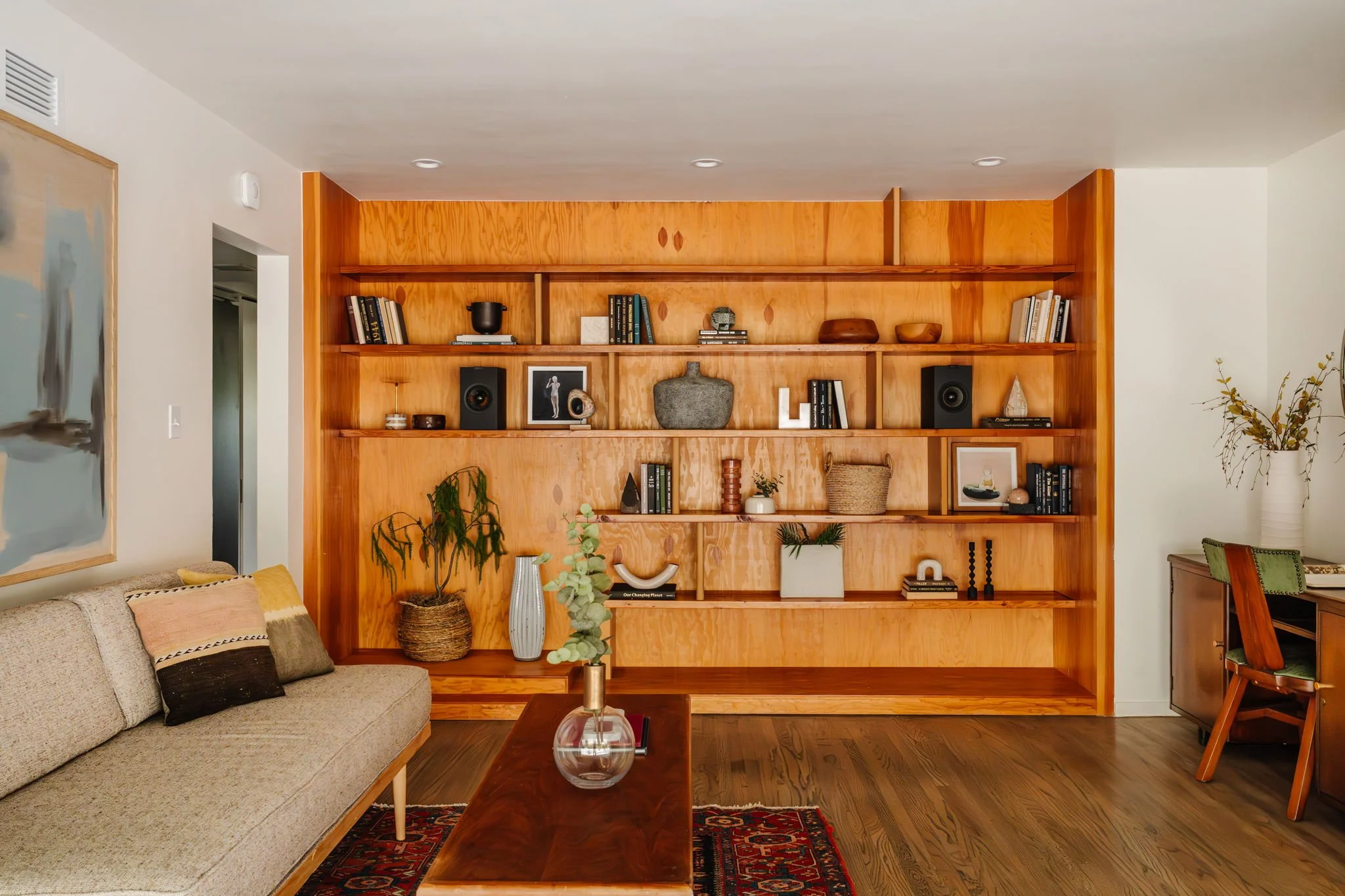  Wide shot of a solid wood built in bookshelf accessorized with books and objects. A MCM daybed is on the left wall, and a desk and chair are on the right wall.  