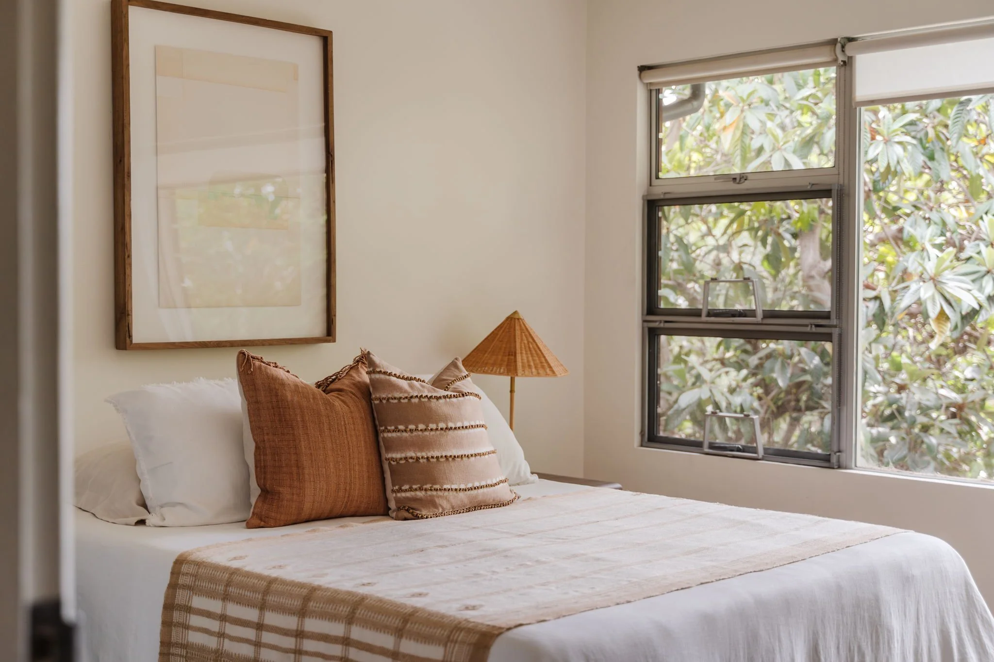  Vignette of bedroom outfitted in neutral colors. Two textured throw pillows accent the bed, a neutral piece of abstract art hangs above it, and a structural rattan lamp sits to the right. 