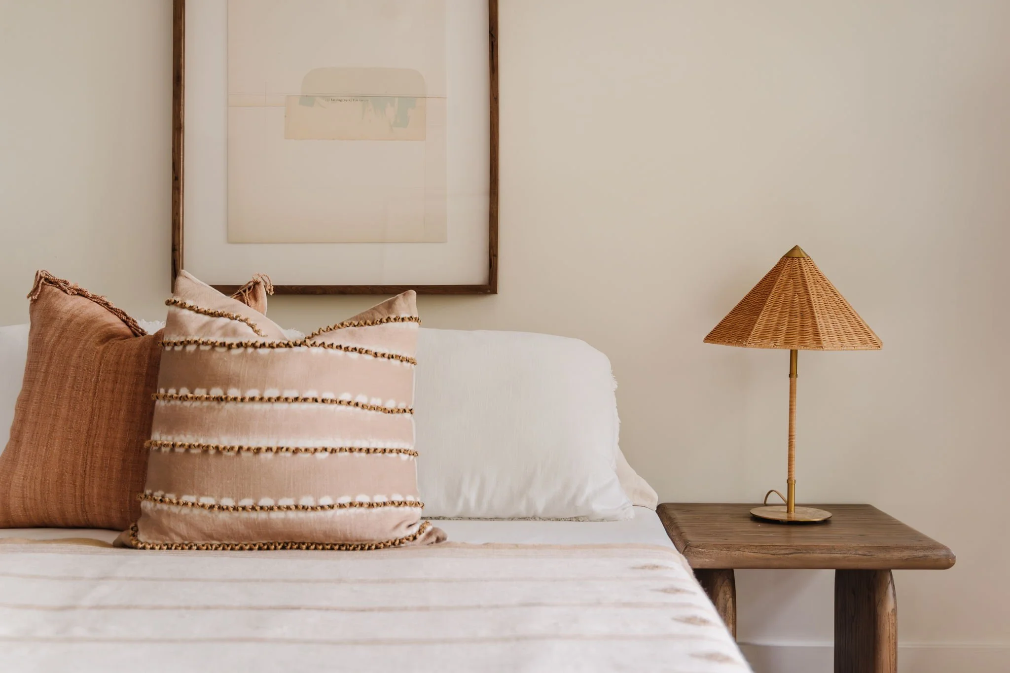  Vignette of bedroom outfitted in neutral colors. Two textured throw pillows accent the bed, a neutral piece of abstract art hangs above it, and a structural rattan lamp sits to the right on a wooden nightstand.  