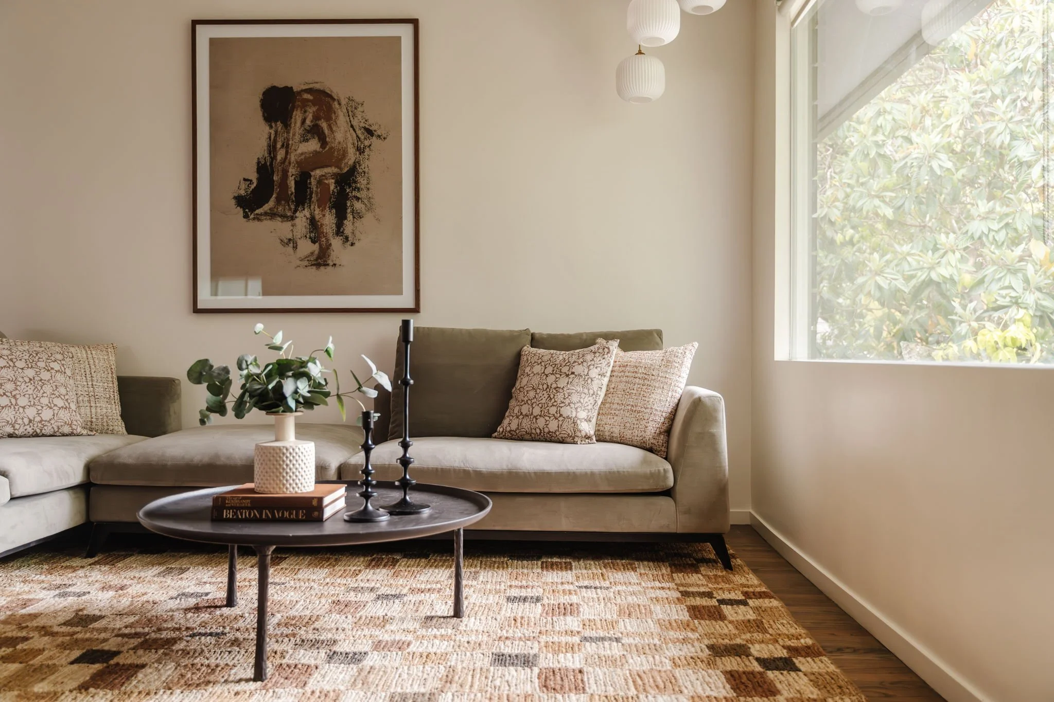  Neutral checkered rug with a sage green velvet couch, round iron-look coffee table, and portrait of a woman.  