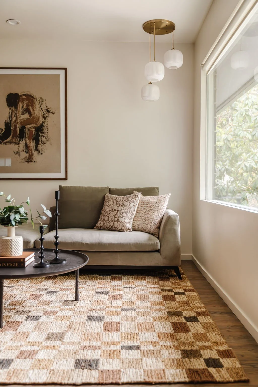  Neutral checkered rug with a sage green velvet couch, round iron-look coffee table, and portrait of a woman.  