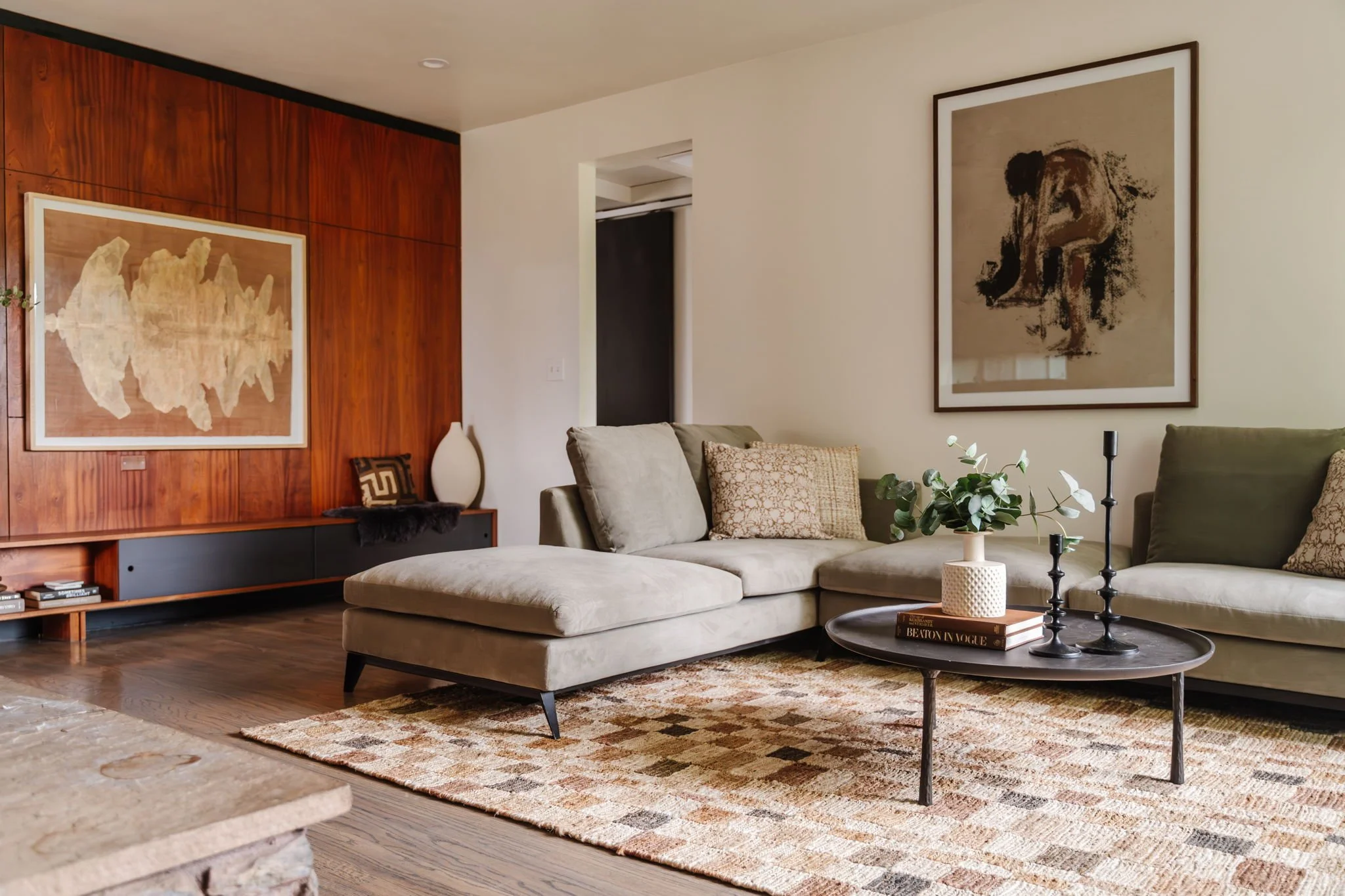  Wide shot of a living room with the left wall clad in wood paneling. A neutral checkered rug with a sage green velvet couch, round iron-look coffee table, and portrait of a woman hung behind the couch.   