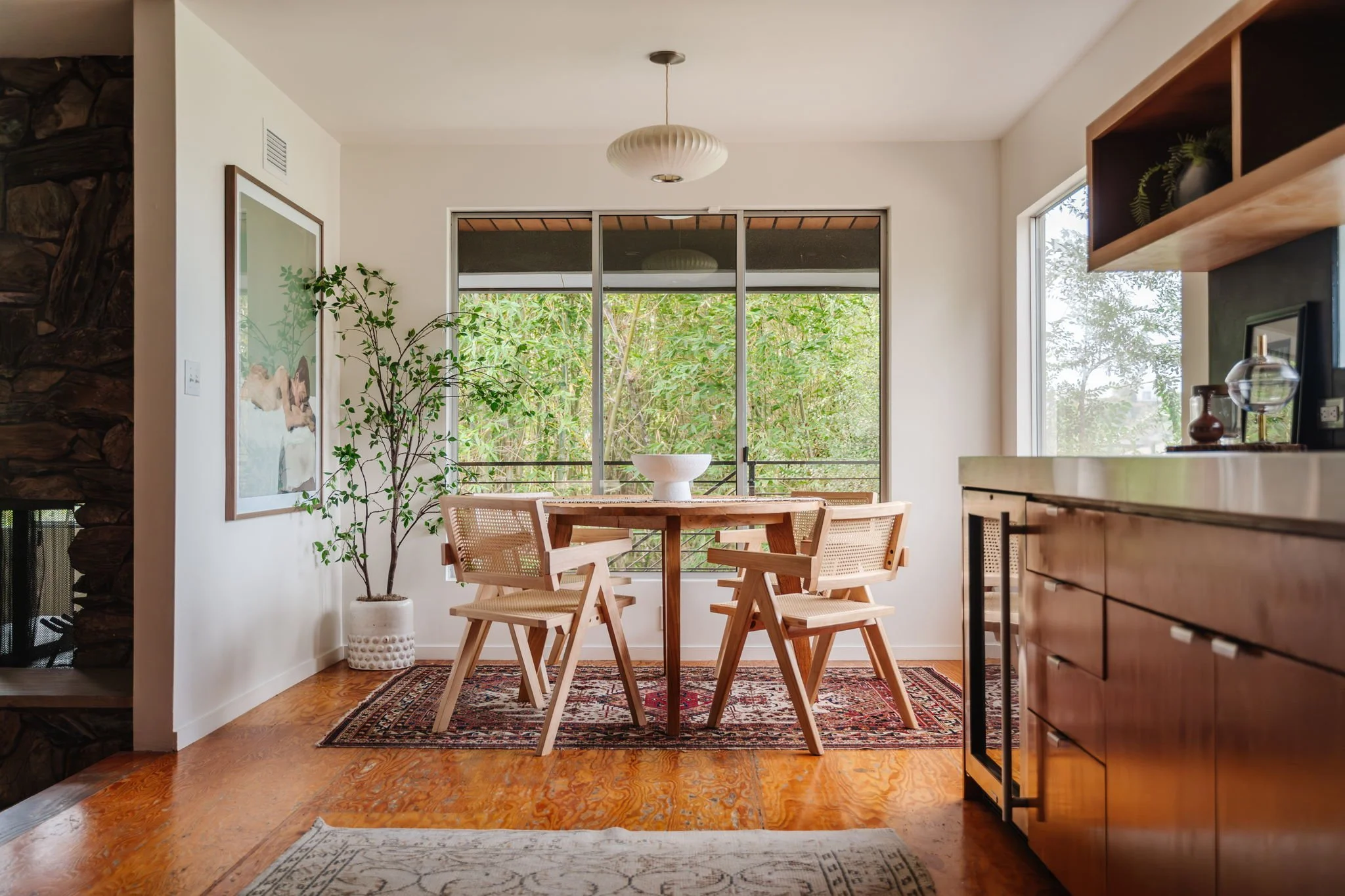  Round rustic wood dining table sitting on a vintage Persian rug, surrounded by four light wood caned dining chairs. A large framed portrait of a woman laying down is hung on the wall to the left. 