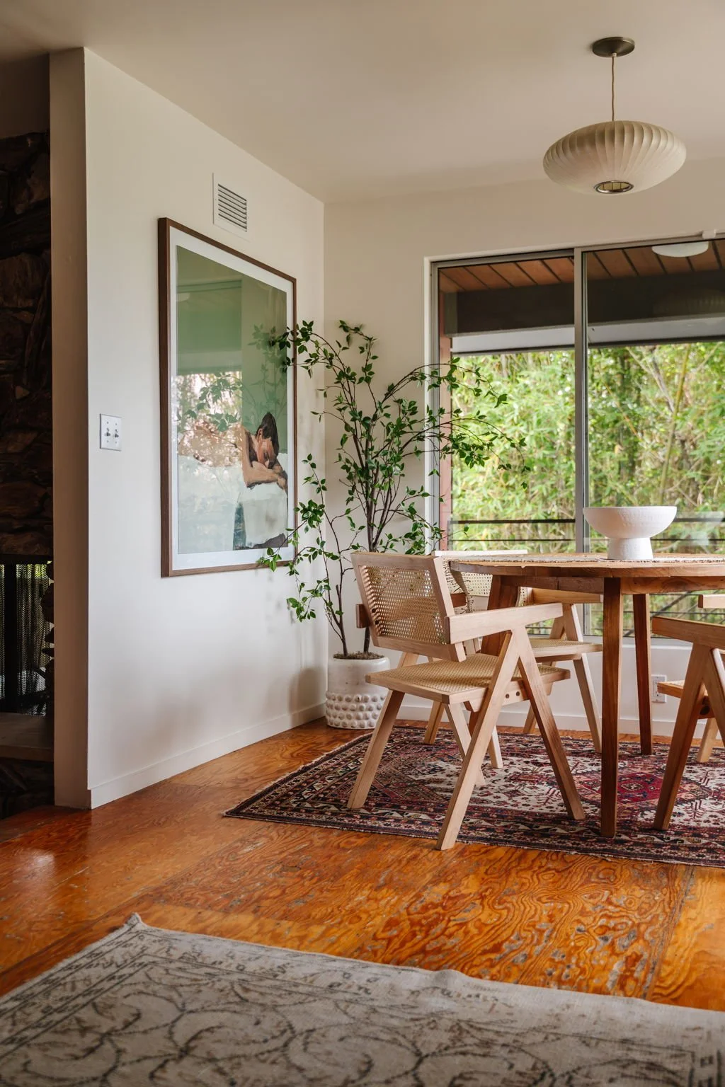  Round rustic wood dining table sitting on a vintage Persian rug, surrounded by four light wood caned dining chairs. A large framed portrait of a woman laying down is hung on the wall to the left. 